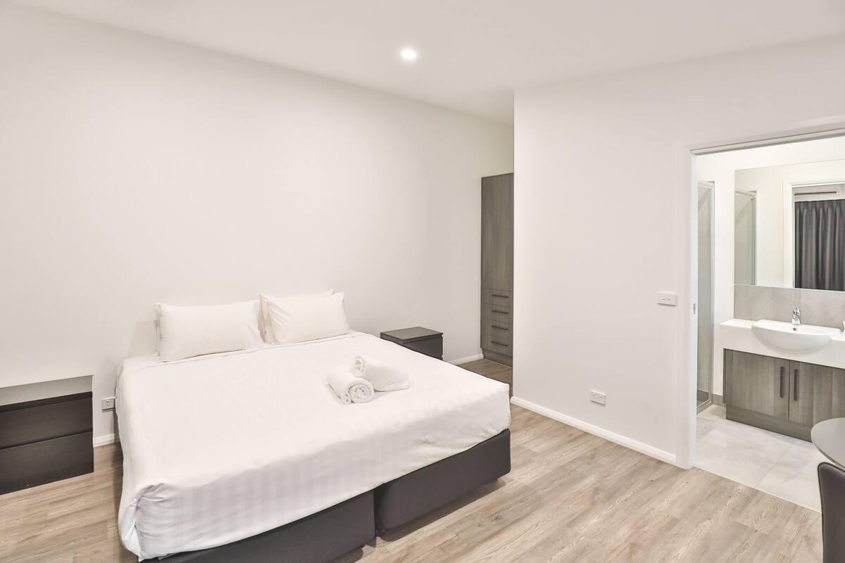 A modern bedroom with a large bed, bedside tables, a built-in wardrobe, and a bathroom with a sink and mirror.