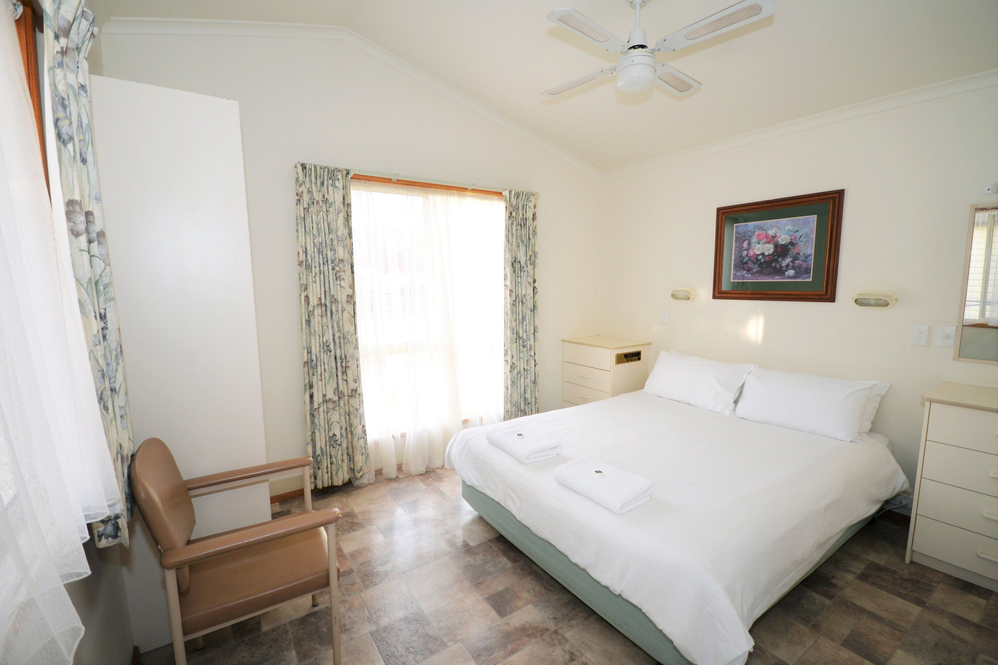 Ein Schlafzimmer mit Bett, Sessel, Nachttisch, Fenster mit Vorhängen und Deckenventilator.