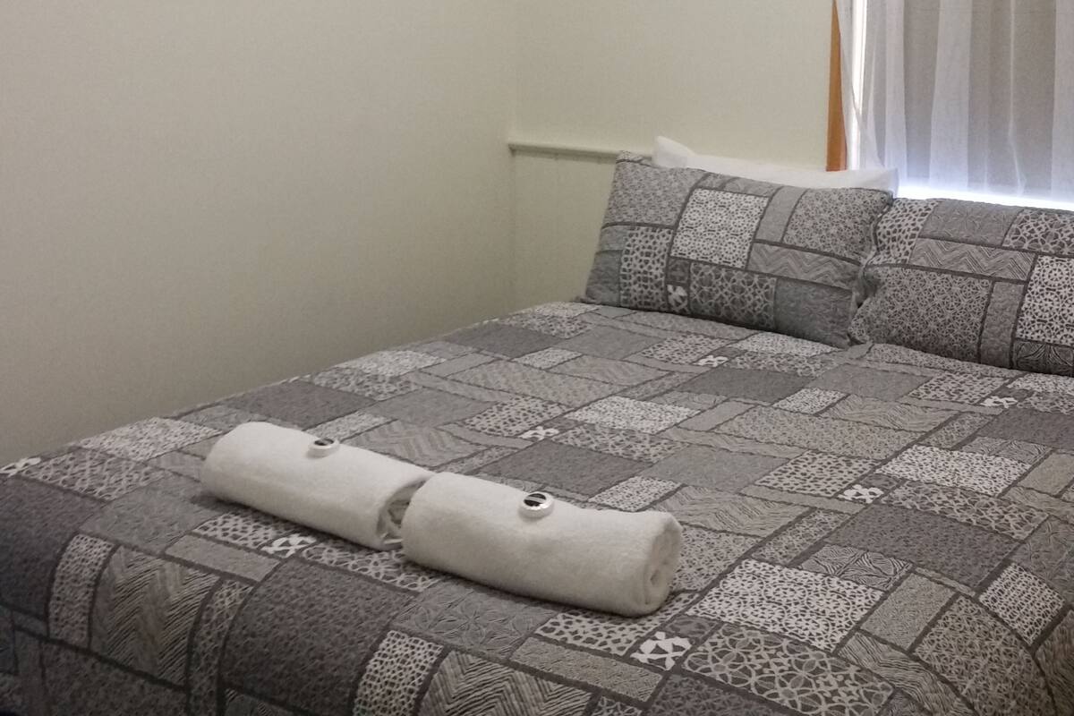 A bedroom with a patterned bedspread, a rolled towel on the bed, and a painting on the wall.
