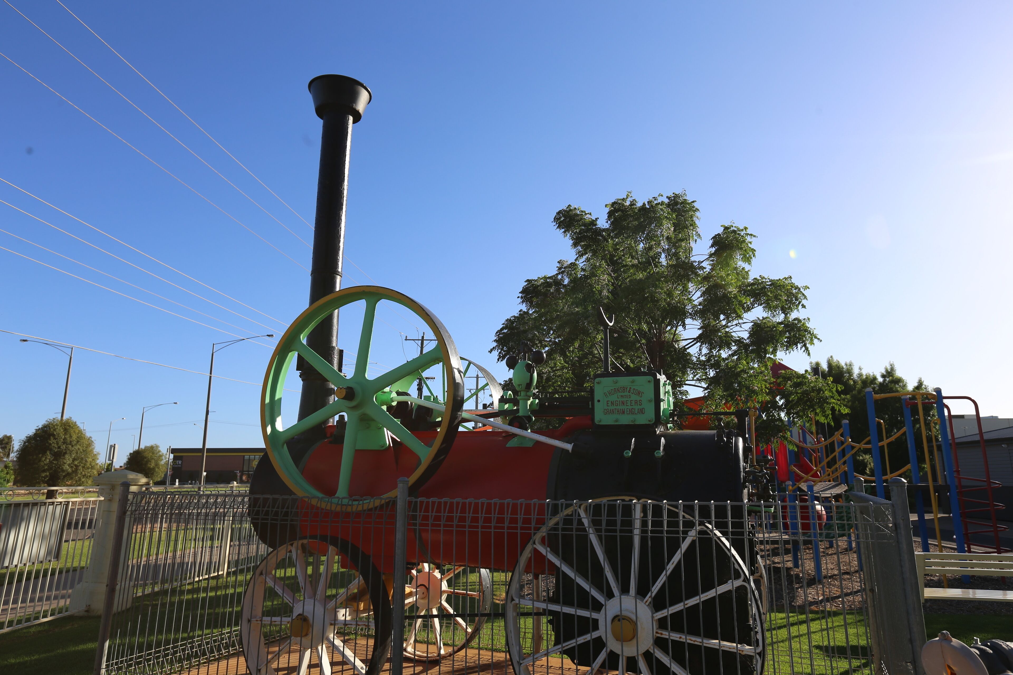 A steam locomotive with green wheels and a black body, surrounded by a fence, with playground equipment in the background.