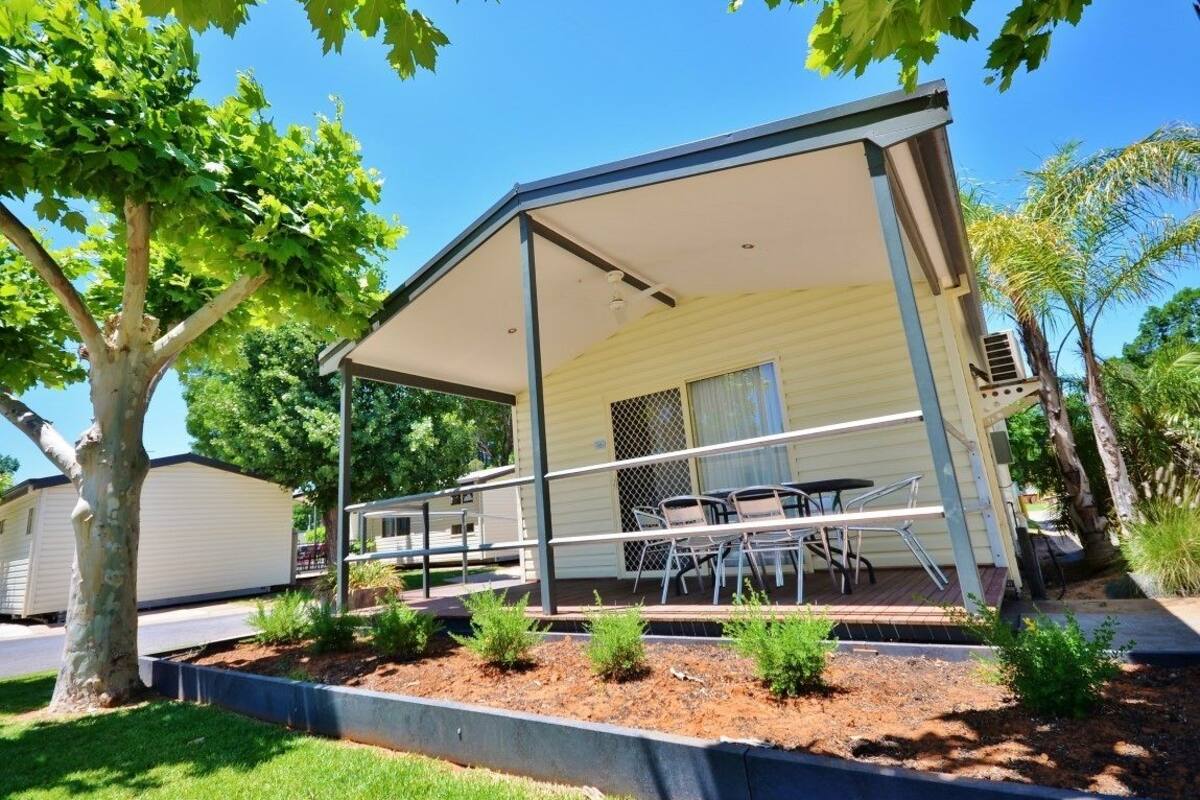 A covered outdoor area with a table and chairs, surrounded by small plants and a tree.