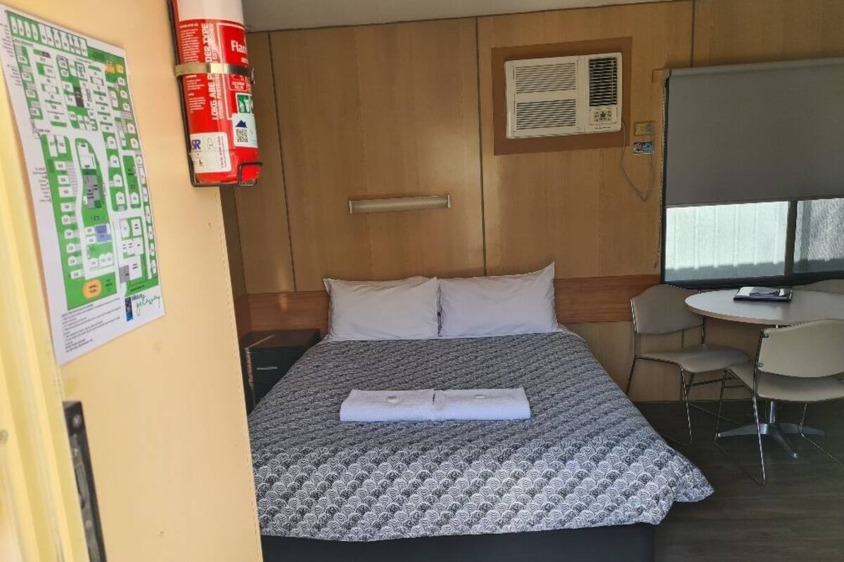 A hotel room with a bed, a desk, a chair, a fire extinguisher, and an air conditioning unit.