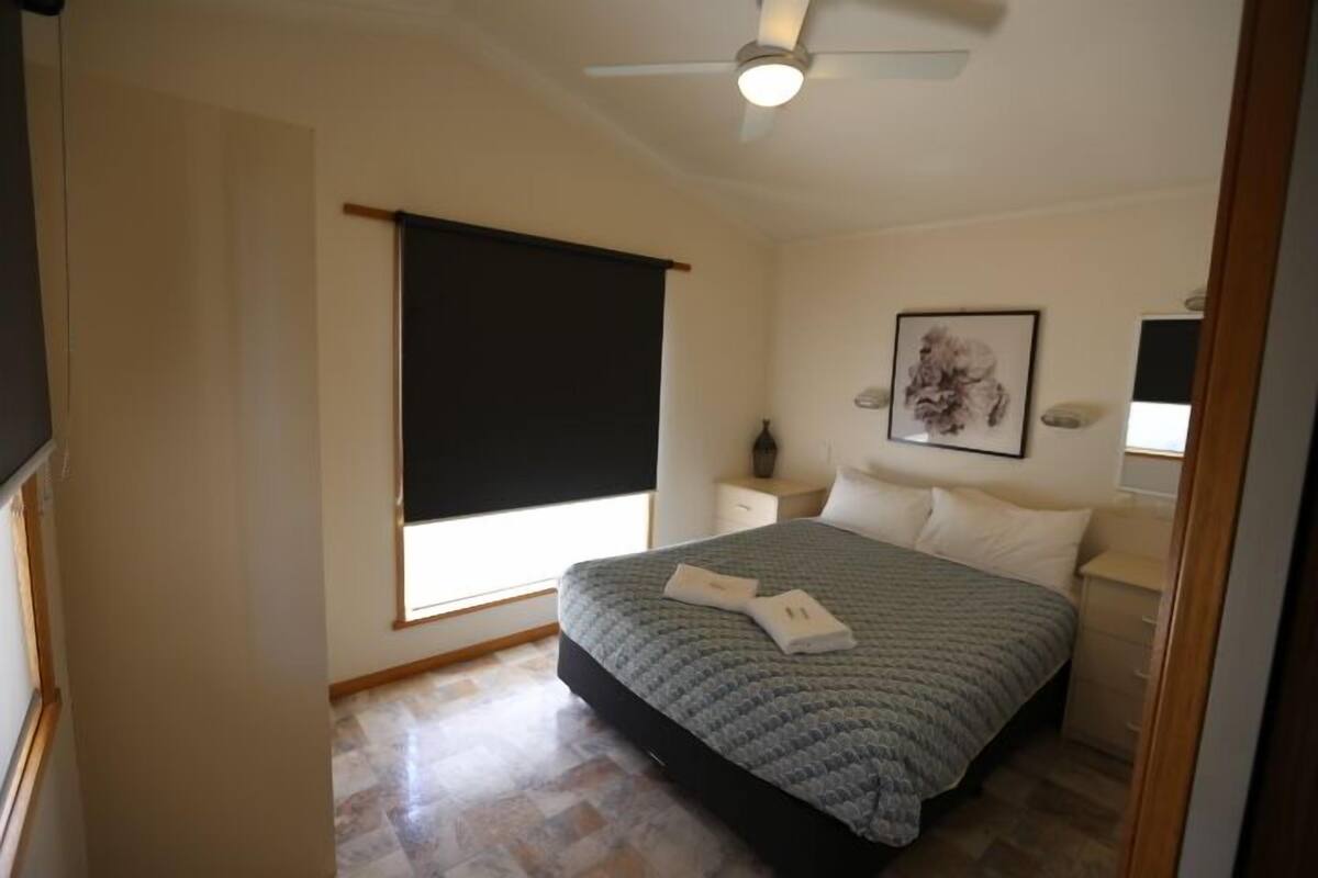 A bedroom with a bed, a nightstand, a ceiling fan, a window with a blind, and a framed picture on the wall.