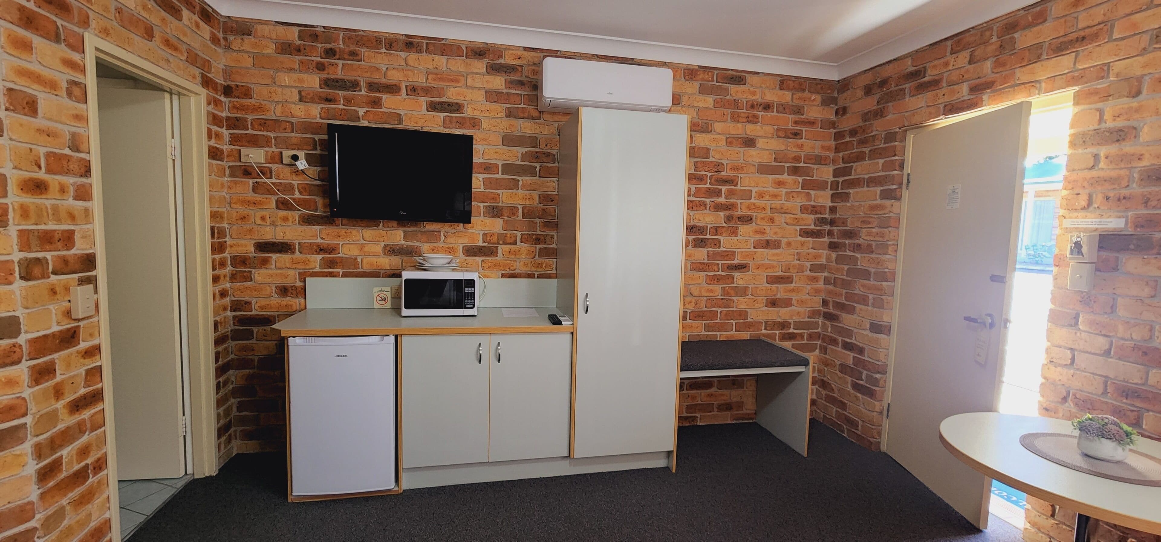 A room with brick walls, a flat-screen TV mounted above a white cabinet, a microwave, and a small refrigerator.