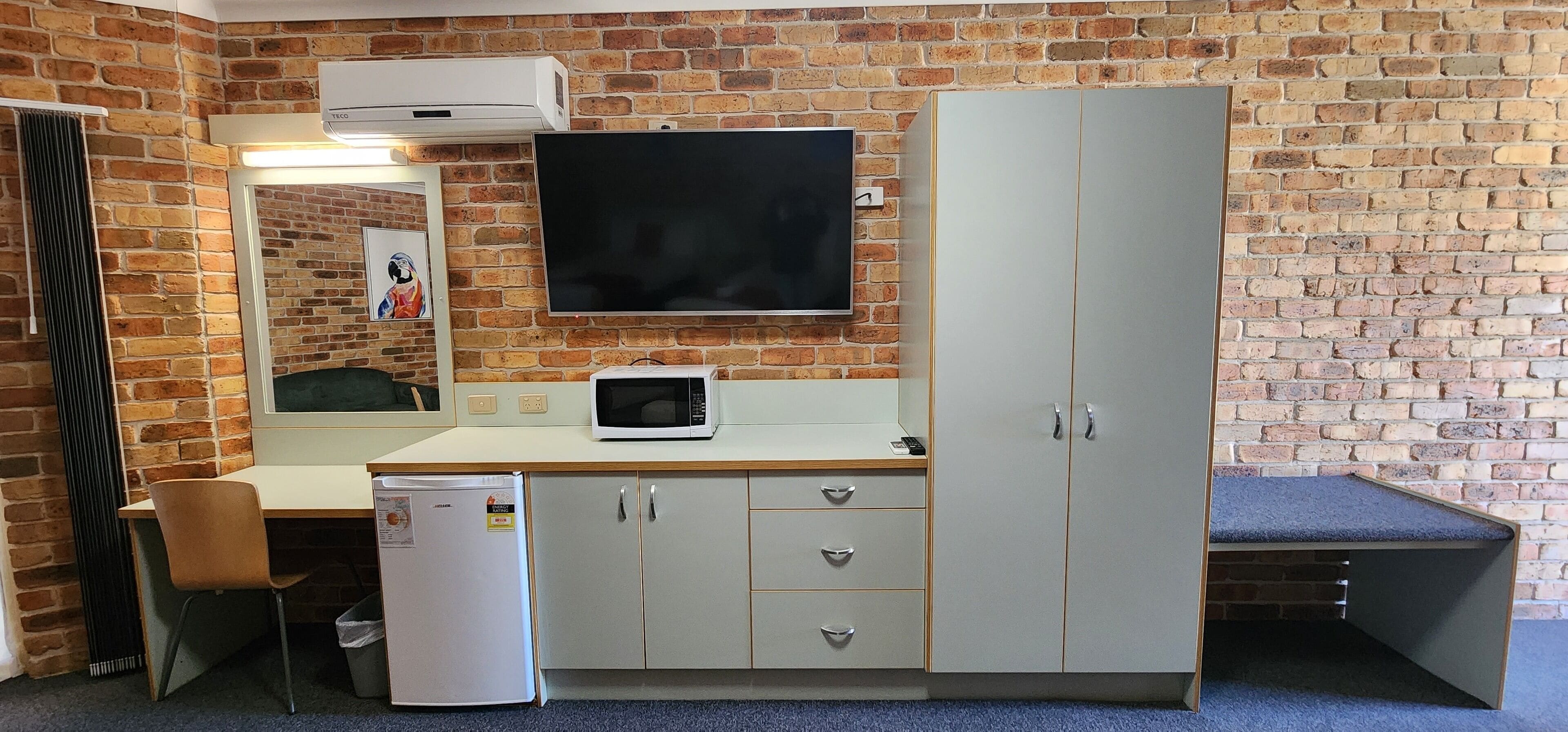 A compact room with a brick wall, a flat-screen TV mounted above a white desk, a microwave, a refrigerator, and a wardrobe with drawers.