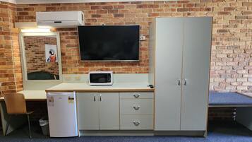 A compact room with a brick wall, a flat-screen TV mounted above a white desk, a microwave, a refrigerator, and a wardrobe with drawers.