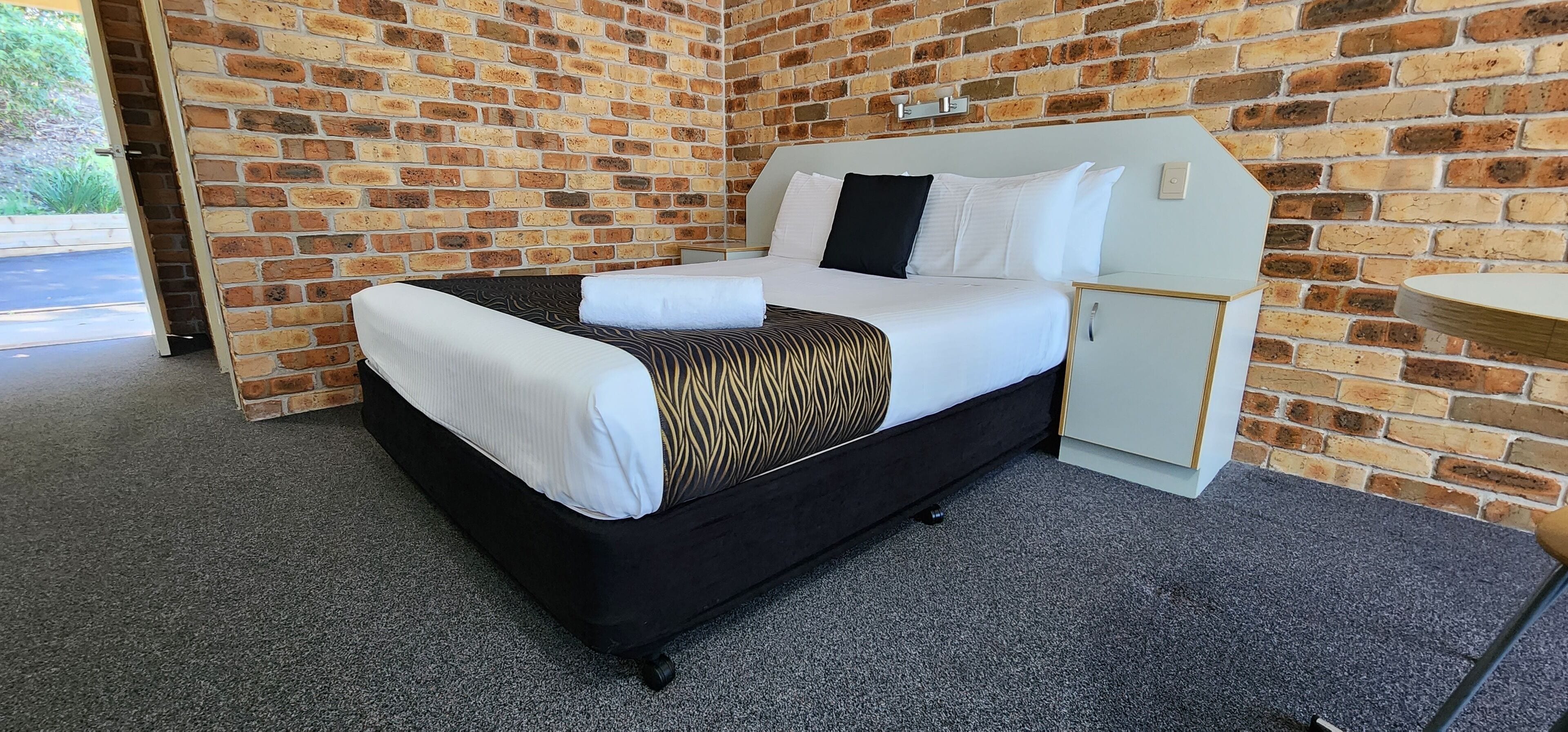 A hotel room with a brick wall, a bed with white and patterned bedding, a small bedside table, and a built-in cabinet.
