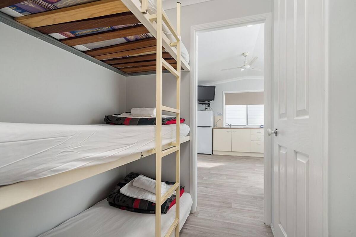 A bunk bed room with a wooden ceiling, a kitchen in the background, and a white door.