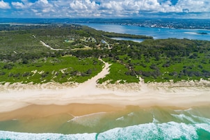 Vista aérea de una zona costera con playa, vegetación y un pueblo distante.