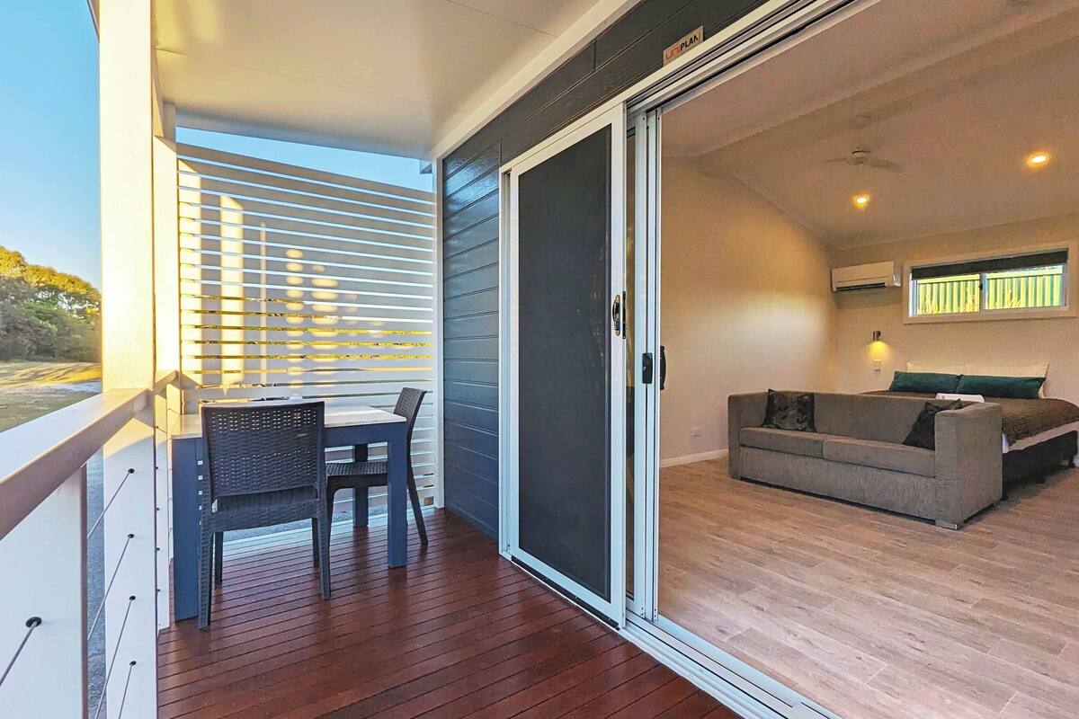 A balcony with a table and chairs, a sliding glass door leading to a living area with a sofa and a bed, and a view of the outdoors.