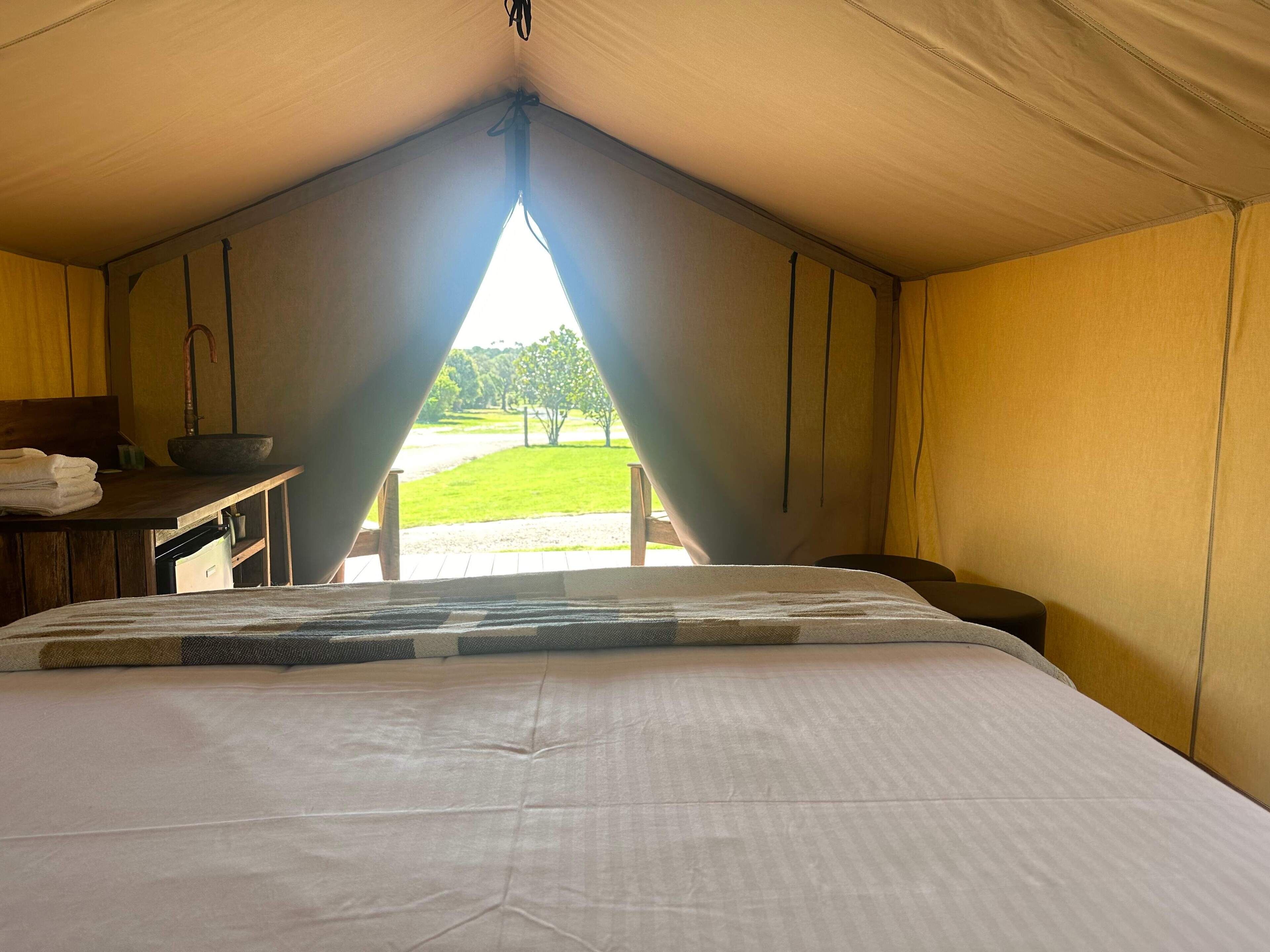 A tented room with a bed, a table with a sink, and a view of a grassy area.