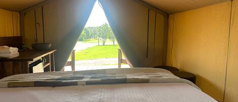 A tented room with a bed, a table with a sink, and a view of a grassy area.