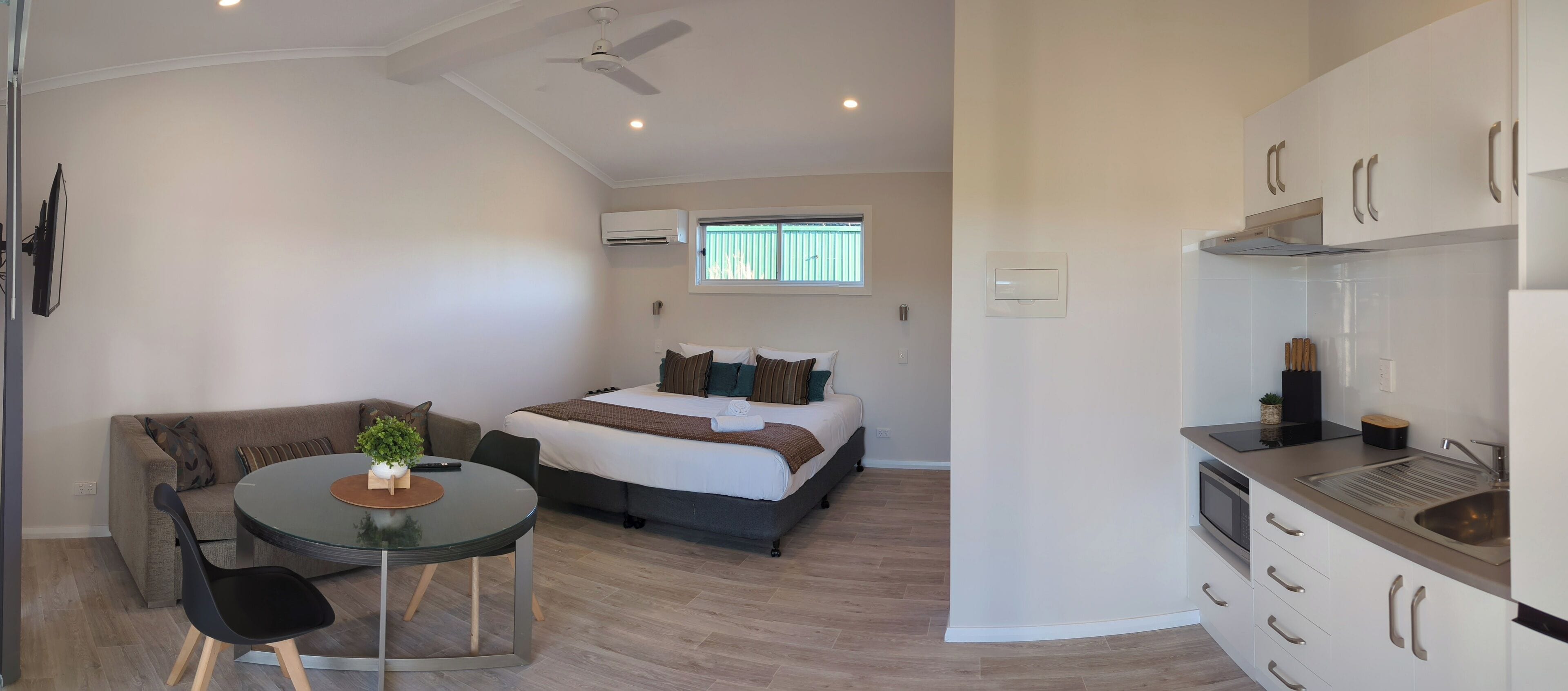 A compact hotel room with a bed, a small dining table with chairs, a kitchenette, and a ceiling fan.