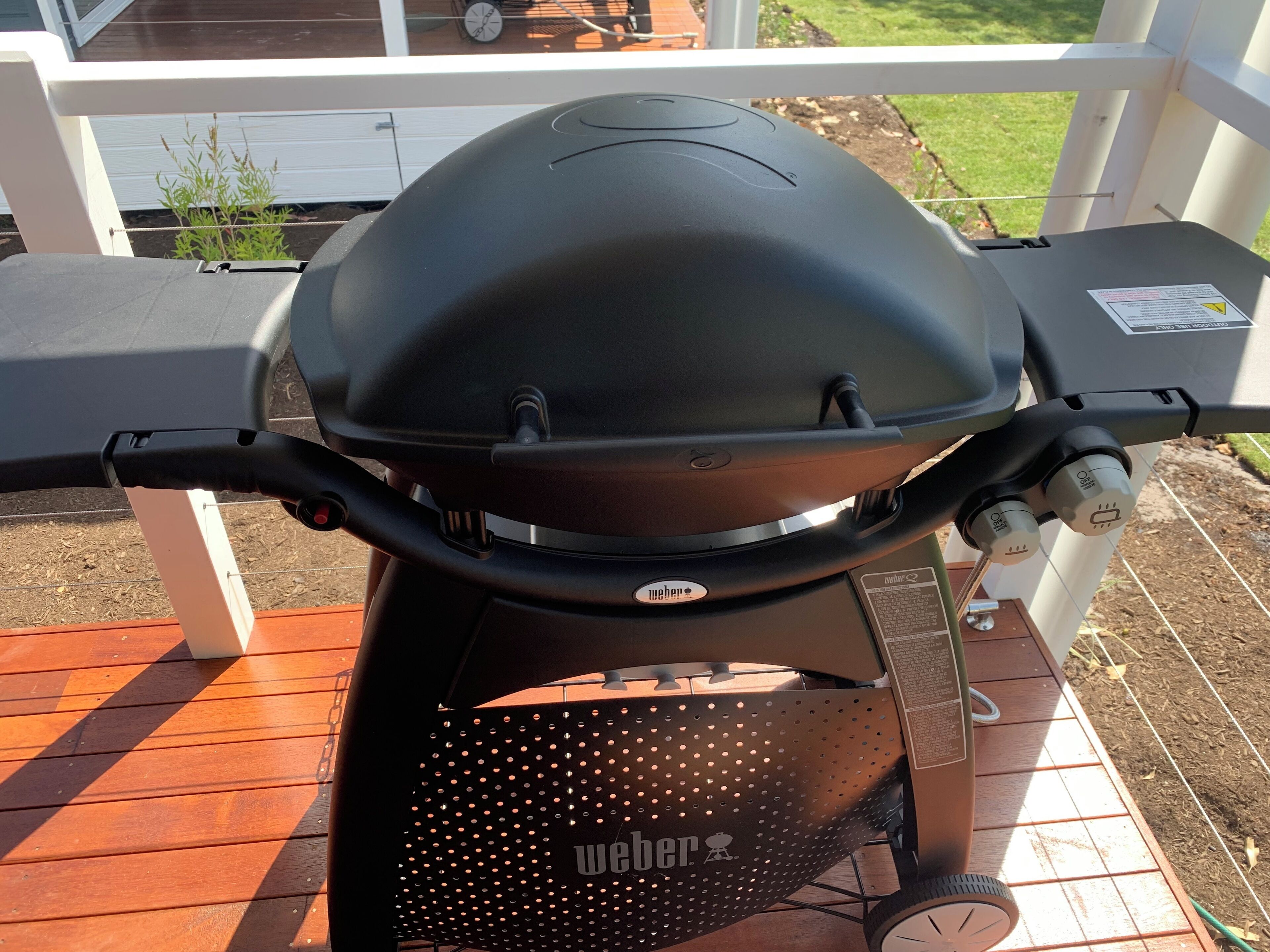 A Weber grill on an outdoor deck with a white railing and a view of a garden.