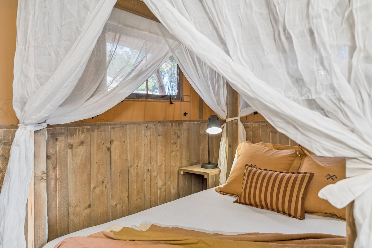 A bedroom with a wooden headboard, a bed with white and brown bedding, a bedside table with a lamp, and a window with a view of trees.