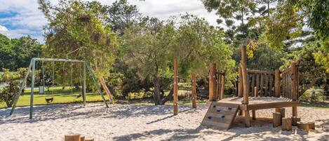 A playground with wooden structures and a swing set in a sandy area surrounded by trees.