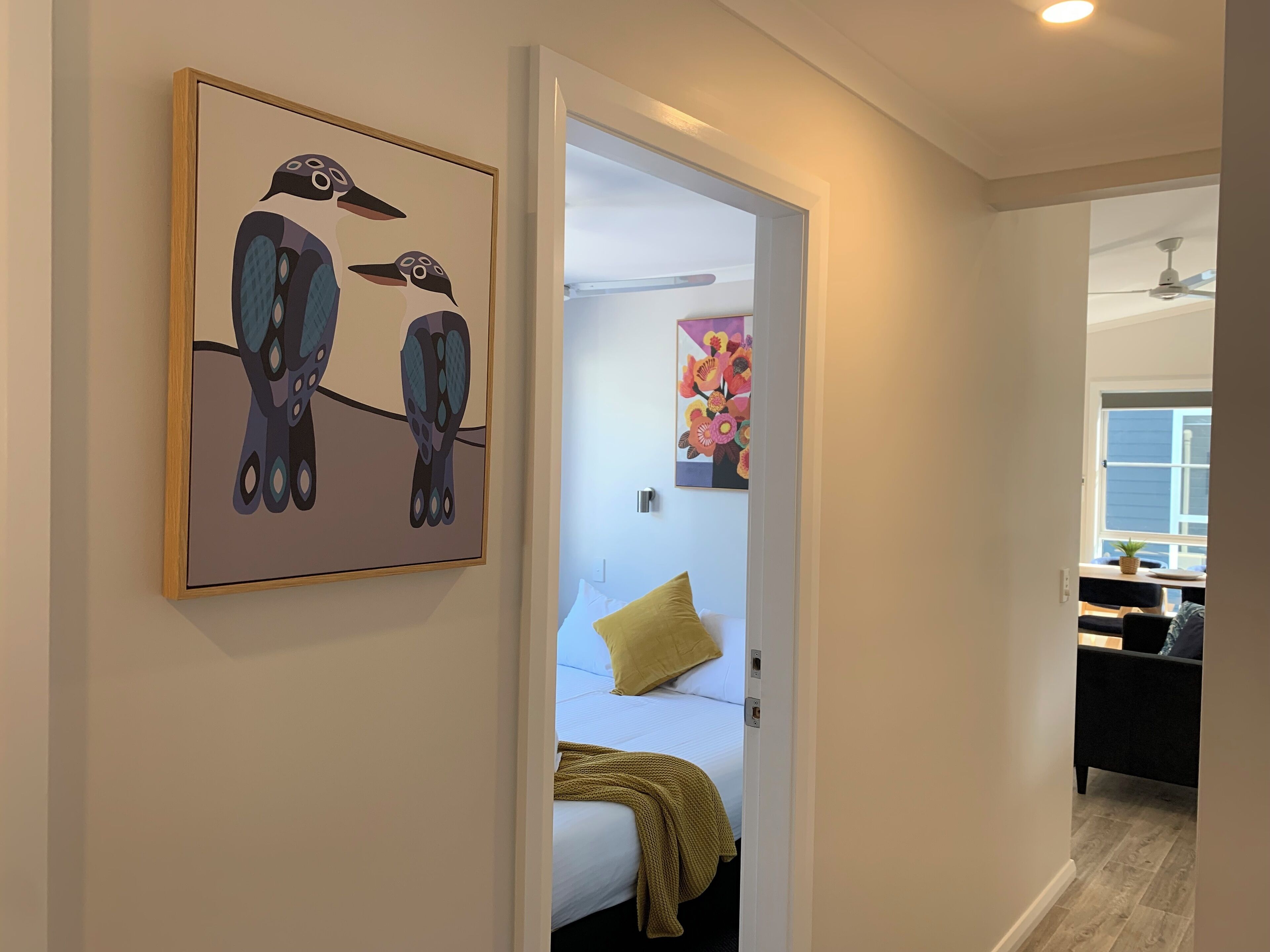 Un pasillo con una cama, un almohadón amarillo y un cuadro de pájaros en la pared.