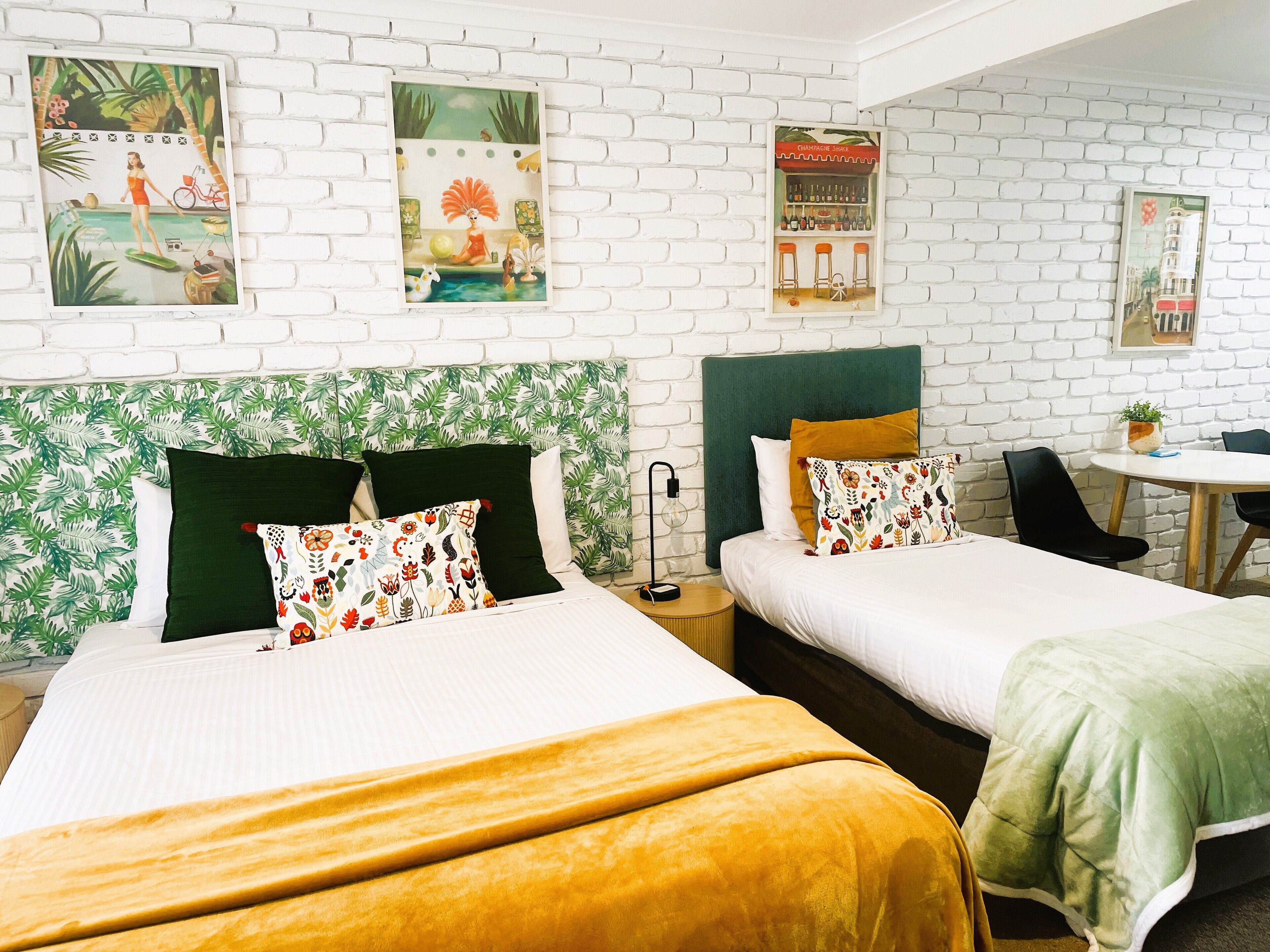 Ein Hotelzimmer mit zwei Betten, einem kleinen Tisch und einem Stuhl. Über den Betten befinden sich Wandbilder und eine weiße Backsteinwand.