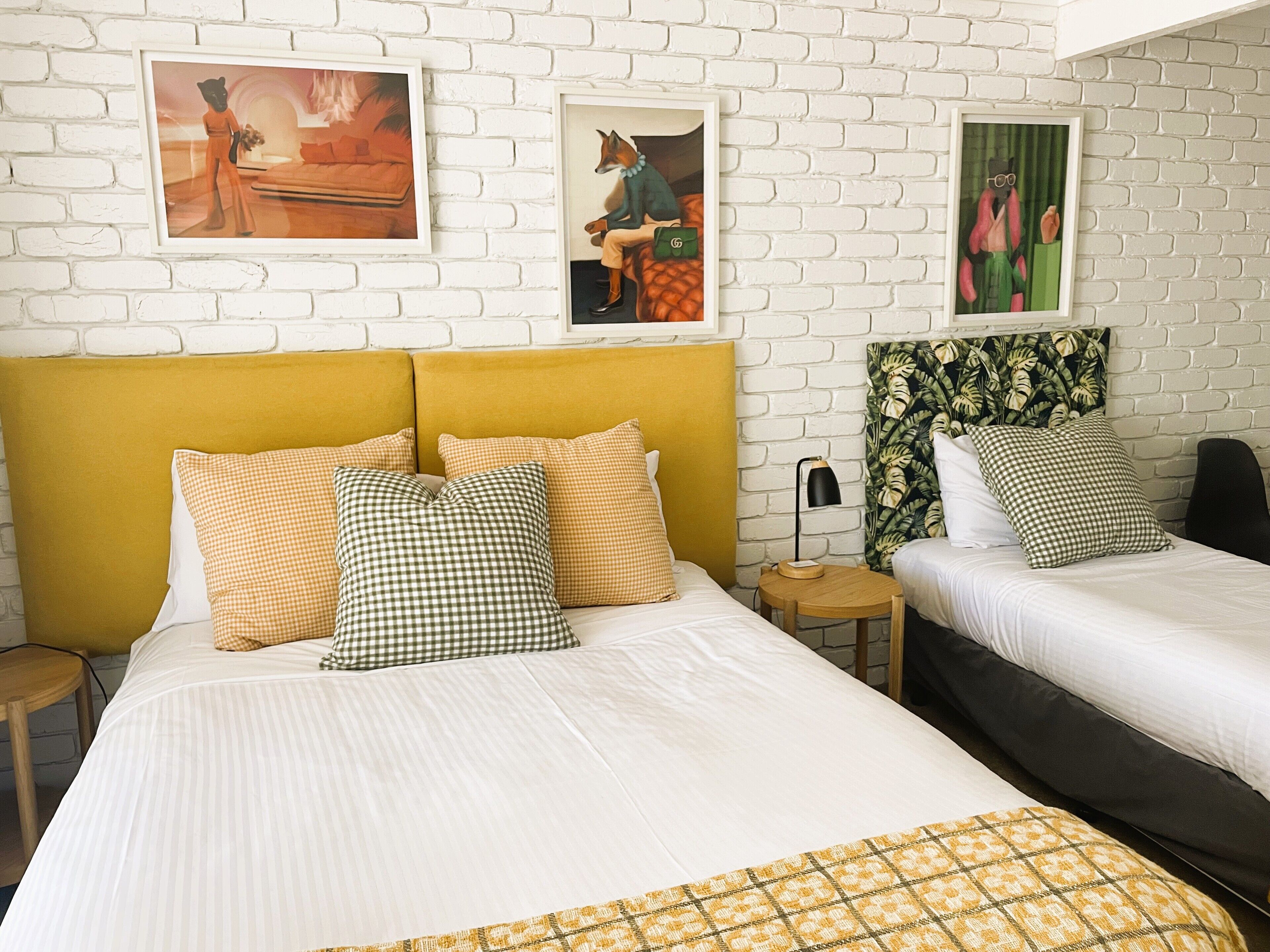 A hotel room with two beds, a yellow headboard, checkered pillows, and artwork on the wall.