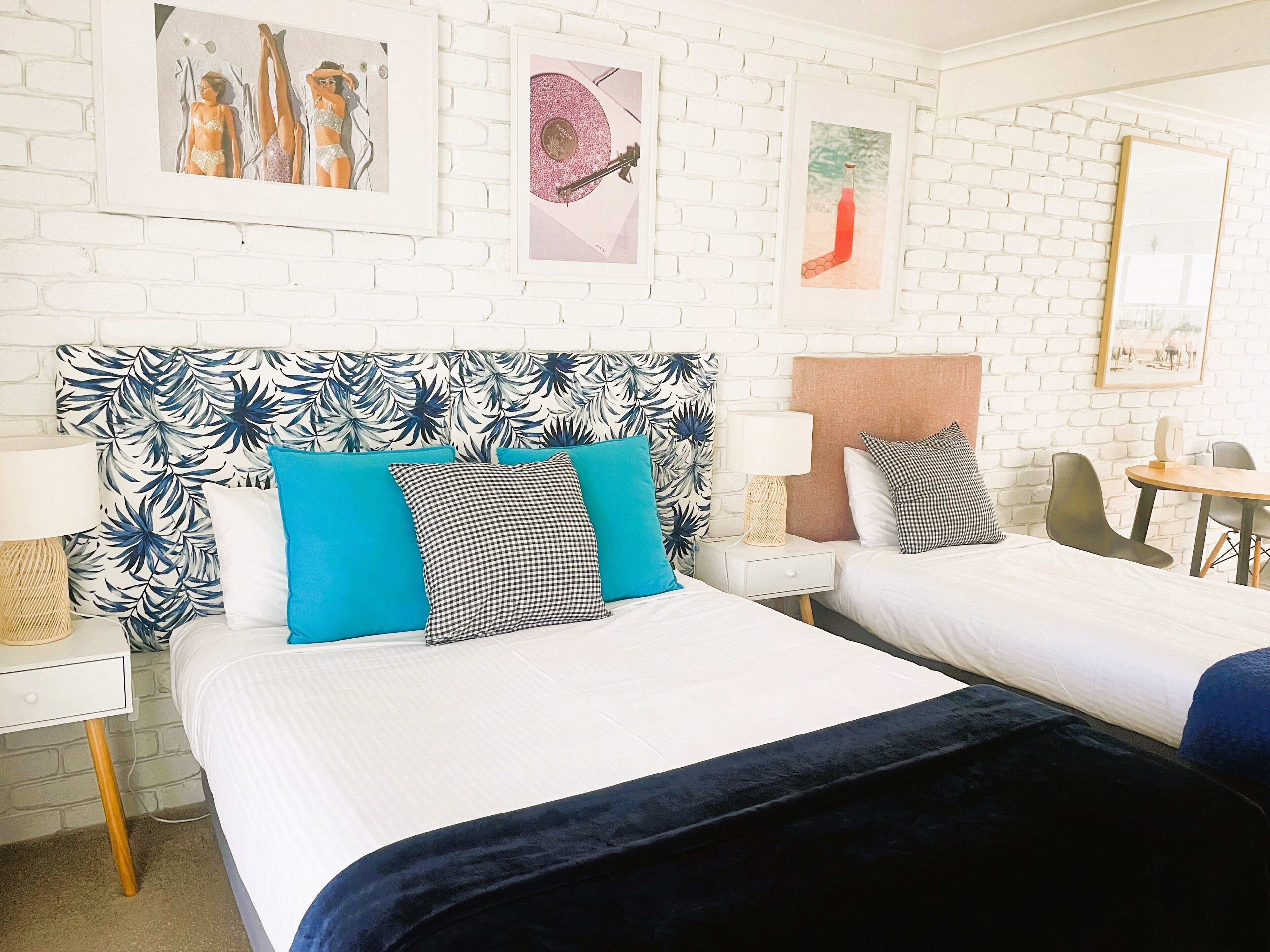 Una habitación de hotel con una pared de ladrillo blanco, dos camas con almohadas a cuadros azules y negros, una mesita con una lámpara y obras enmarcadas en la pared.