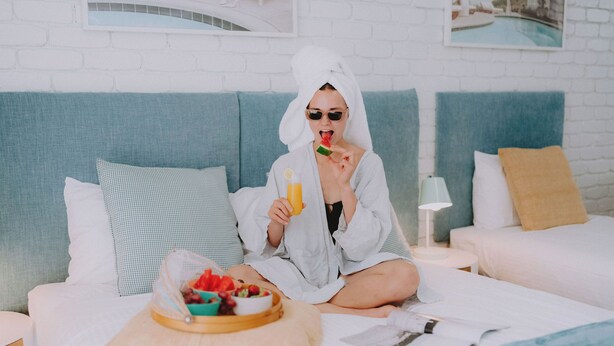 A person sitting on a bed with a tray of fruit and a glass of orange juice, a pillow, and a nightstand with a lamp.