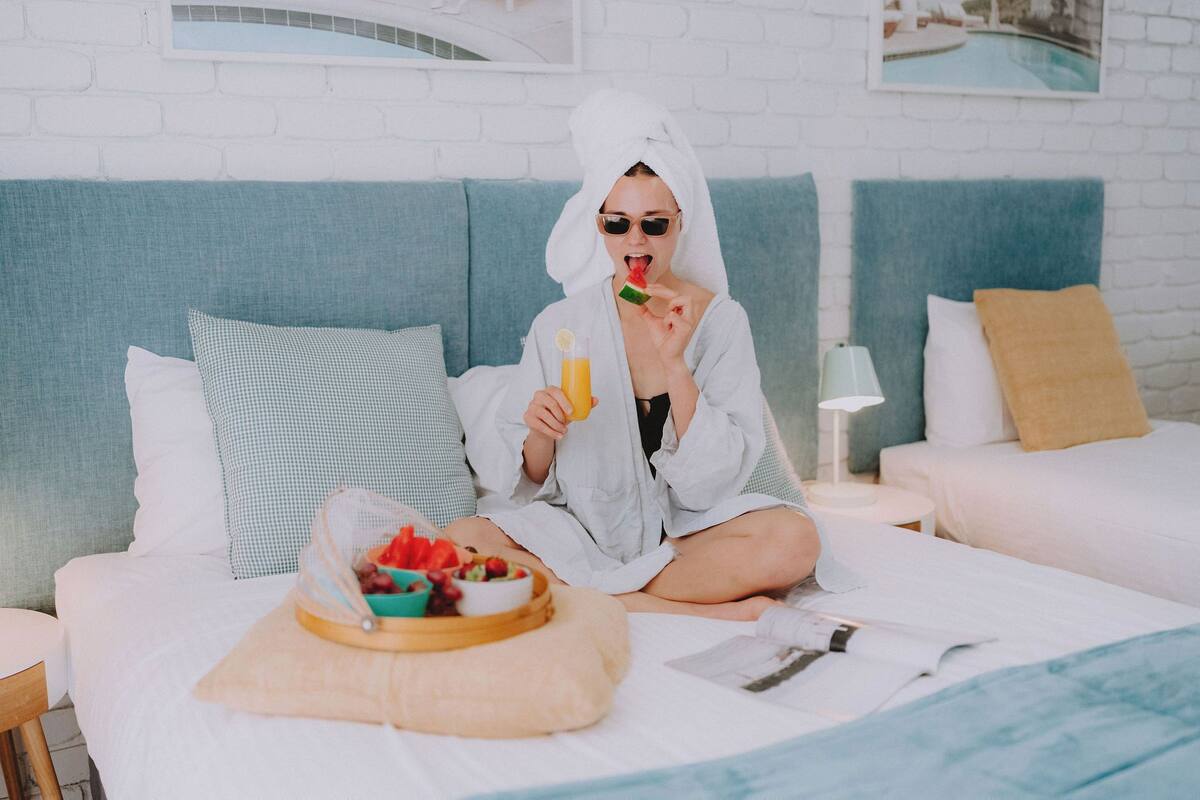 A person sitting on a bed with a tray of fruit and a glass of orange juice, a pillow, and a nightstand with a lamp.