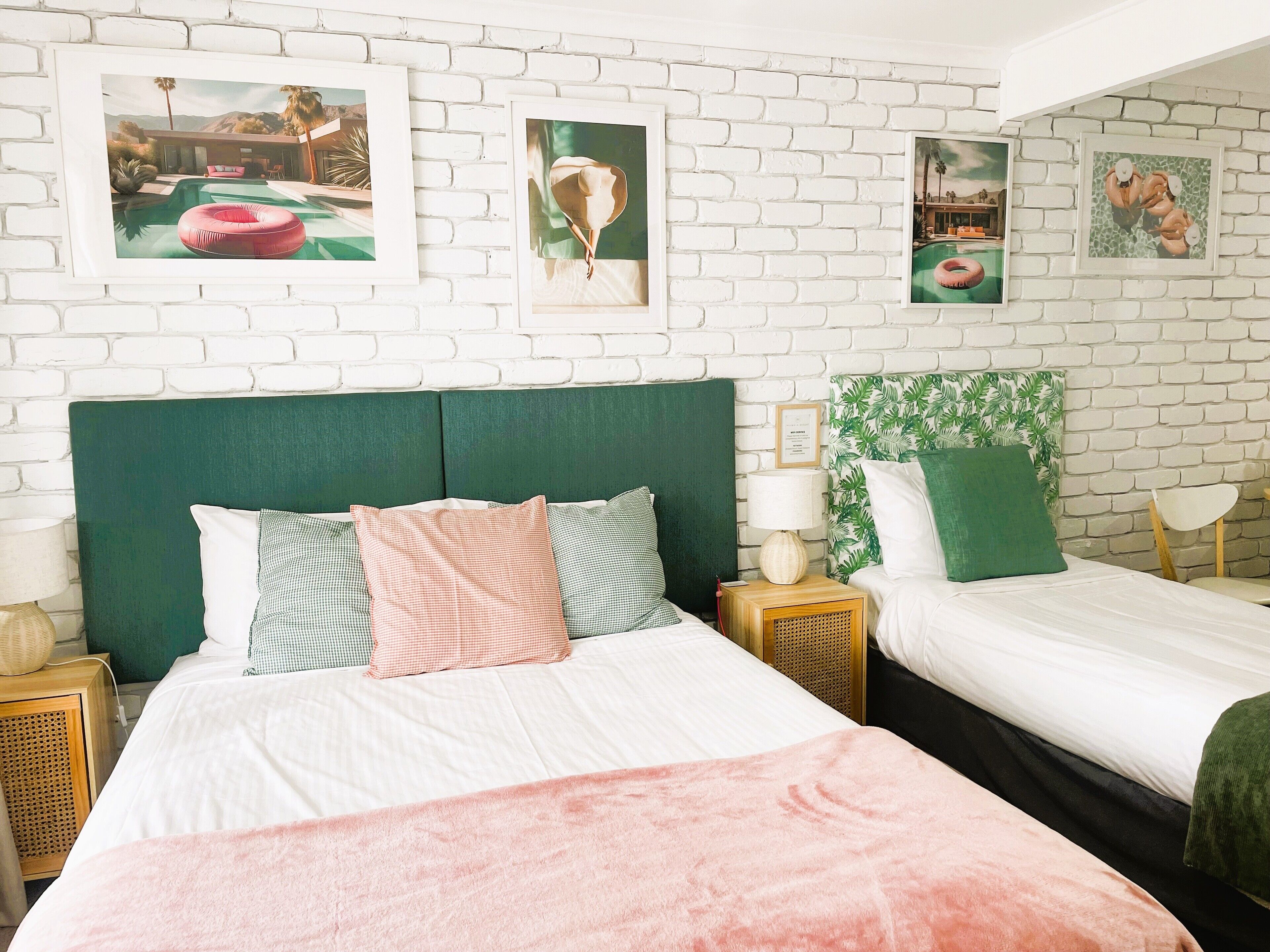 A bedroom with two beds, a green headboard, and a white brick wall with framed pictures.