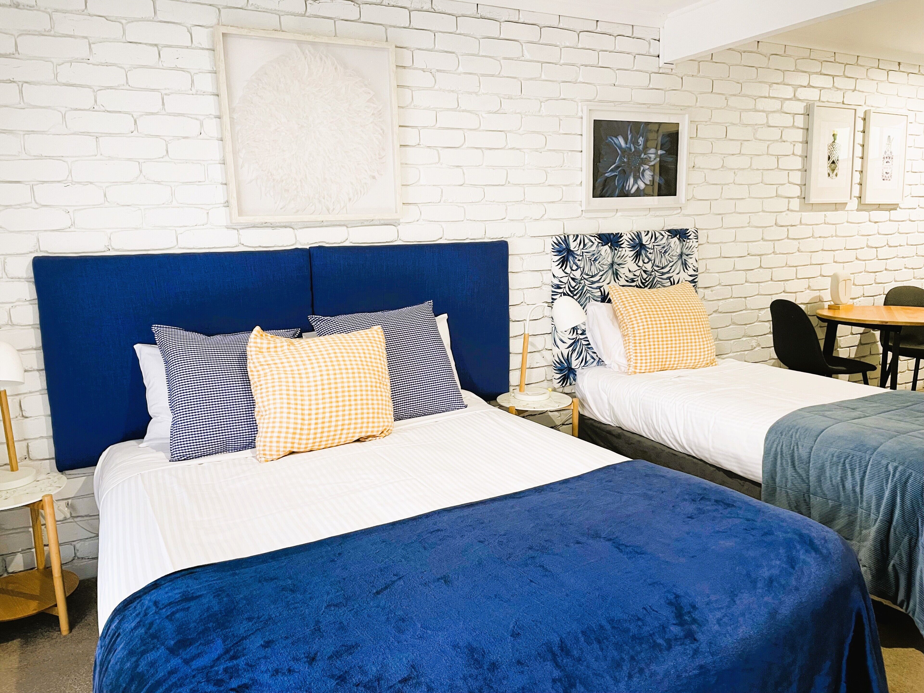 A hotel room with two beds, a white brick wall, and a small table with a lamp.