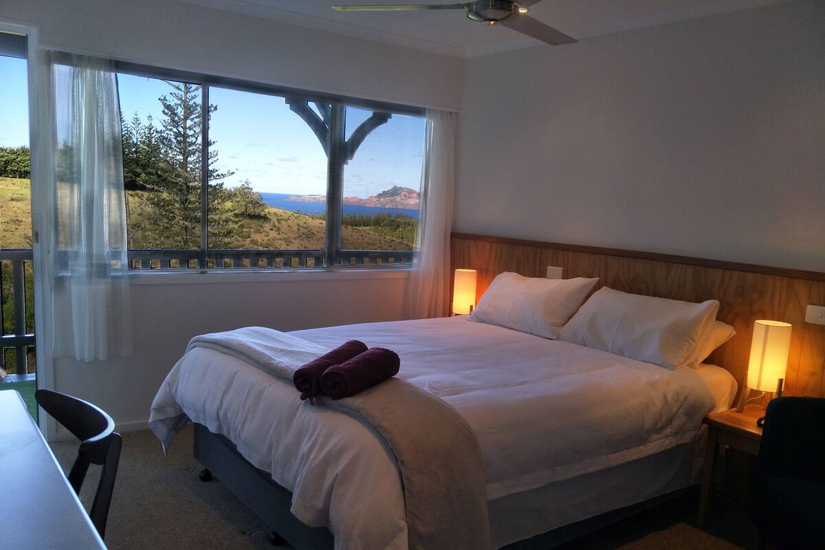 A hotel room with a bed, two bedside lamps, a desk, and a view of a landscape through a window.