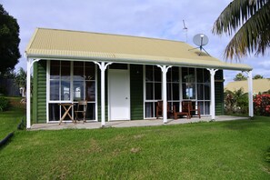 A two-story house with a green exterior and a beige roof, featuring a covered porch with two wooden chairs and a small table.