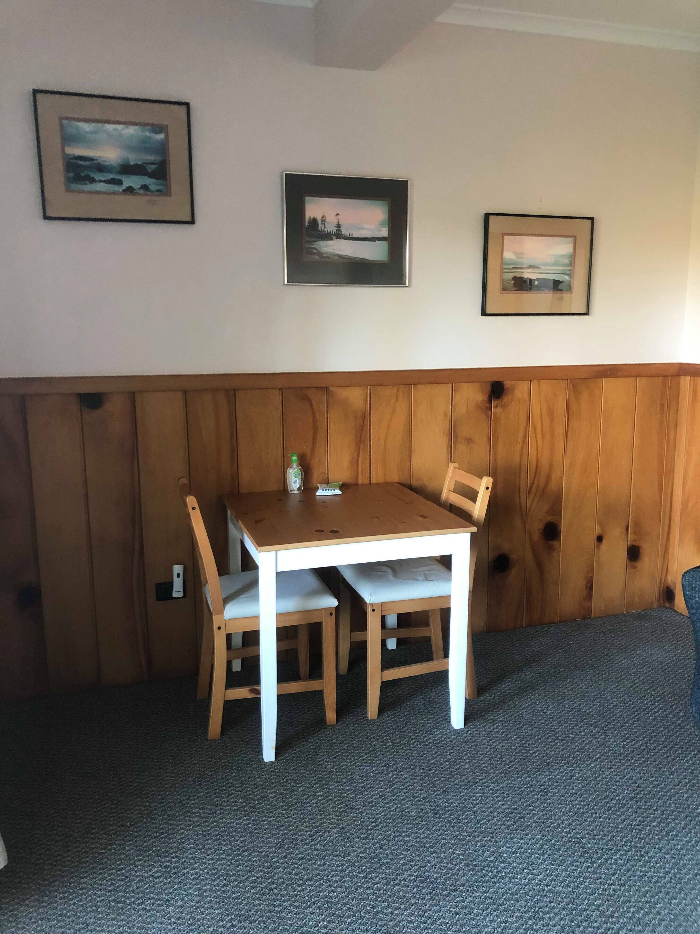 A small wooden table with two chairs, a framed landscape painting, and a wooden panel wall.