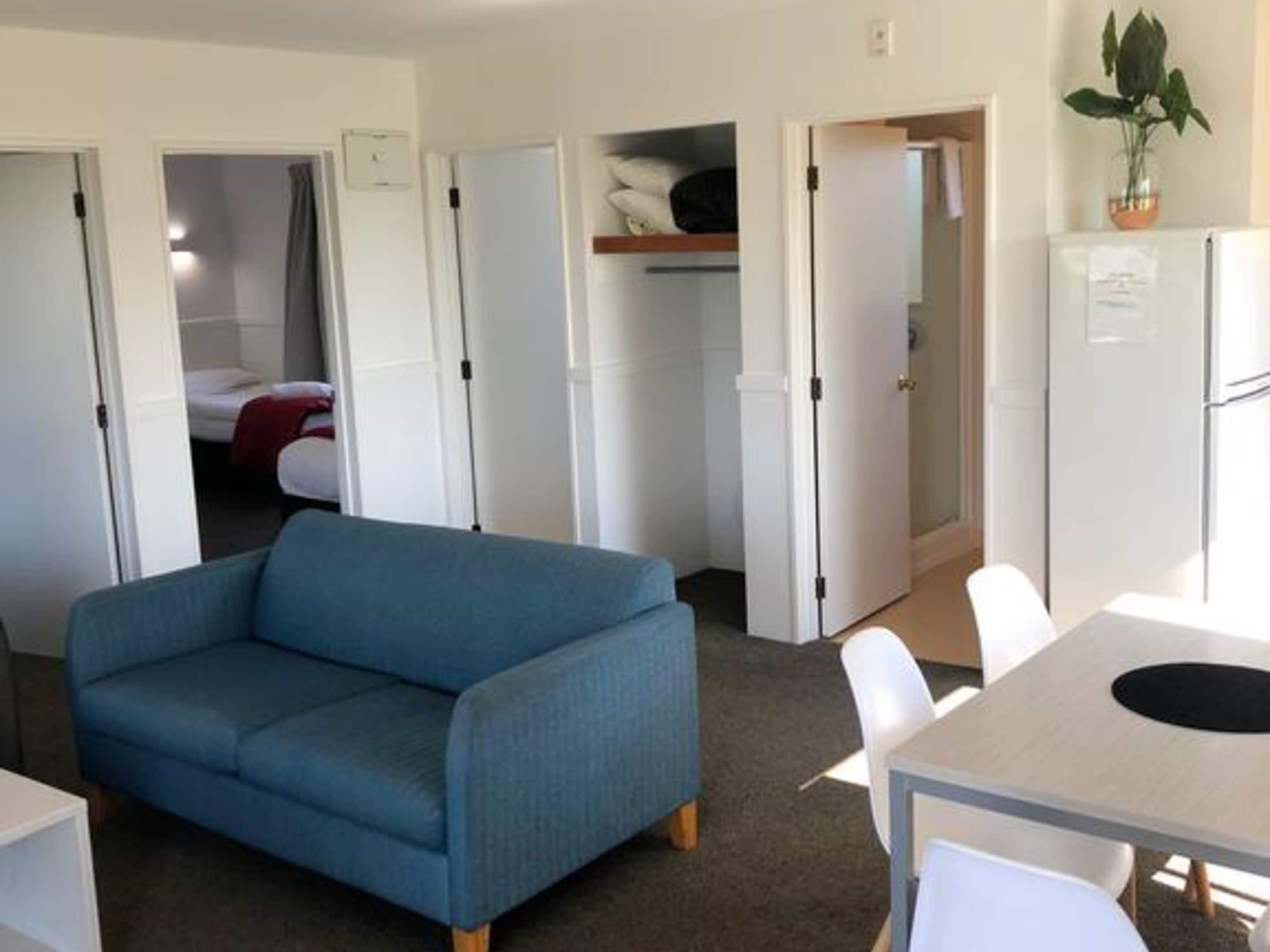 Una habitación de hotel compacta con un sofá azul, una pequeña mesa de comedor y un refrigerador.