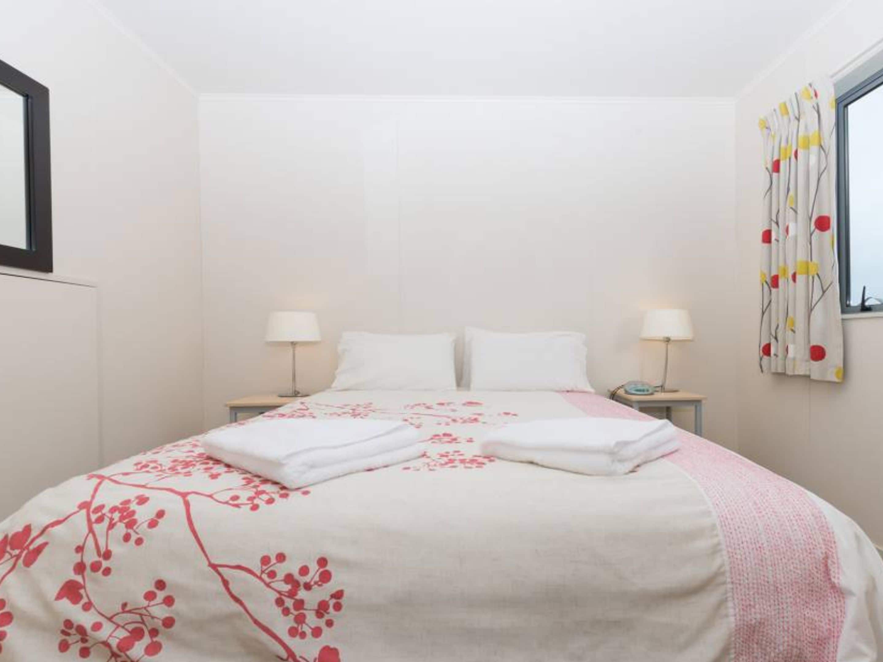 A neatly made bed with white linens and a floral-patterned blanket, two bedside tables with lamps, and a window with a curtain featuring a tree and sun design.
