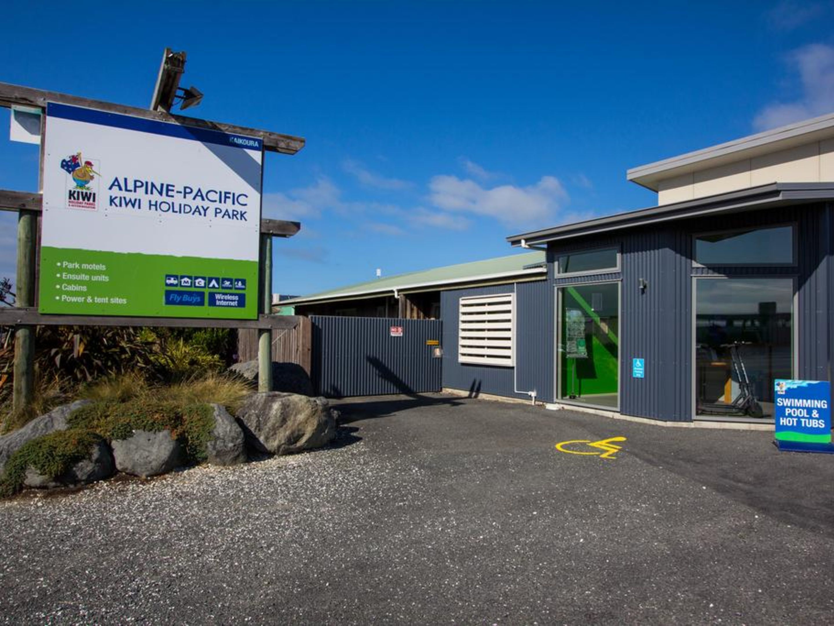 Alpine-Pacific Kiwi Holiday Park entrance with a signboard, a swimming pool, and a building with a green door.