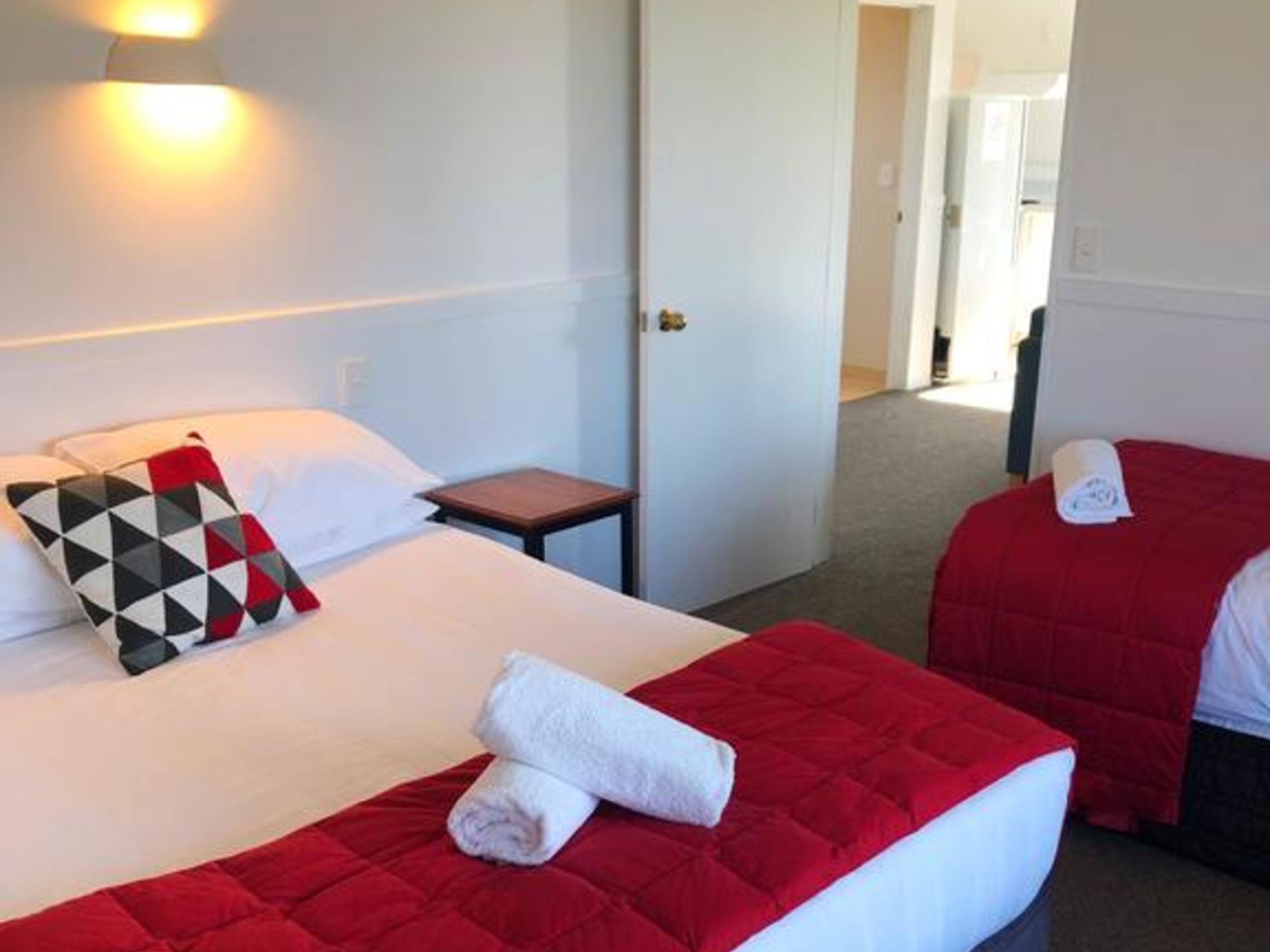 Habitación de hotel con dos camas, cada una con ropa de cama roja y almohadas blancas. Hay una mesita de noche de madera entre las camas, y una pared blanca con una lámpara sobre la cama.