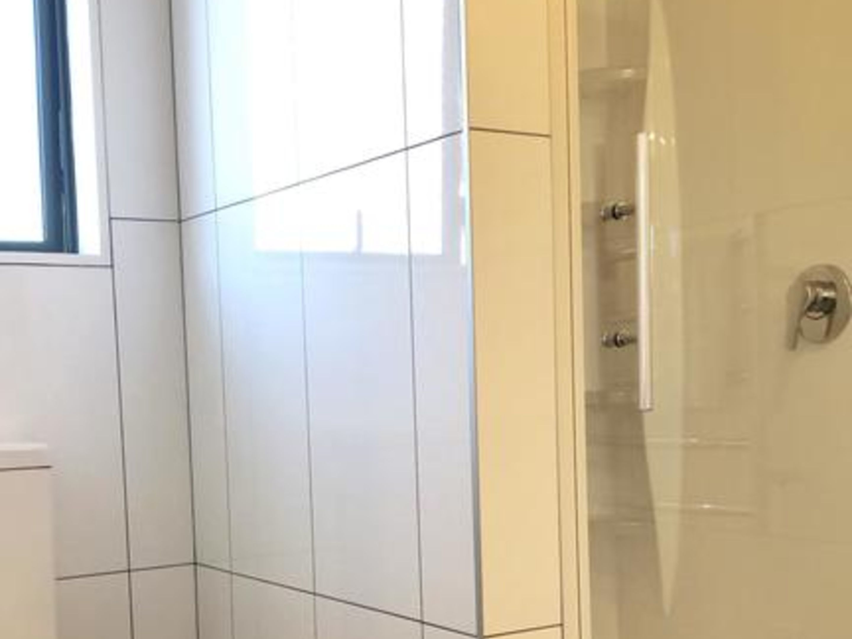 Un baño con azulejos blancos, cabina de ducha con mampara de vidrio y ventana con cortinas azules.