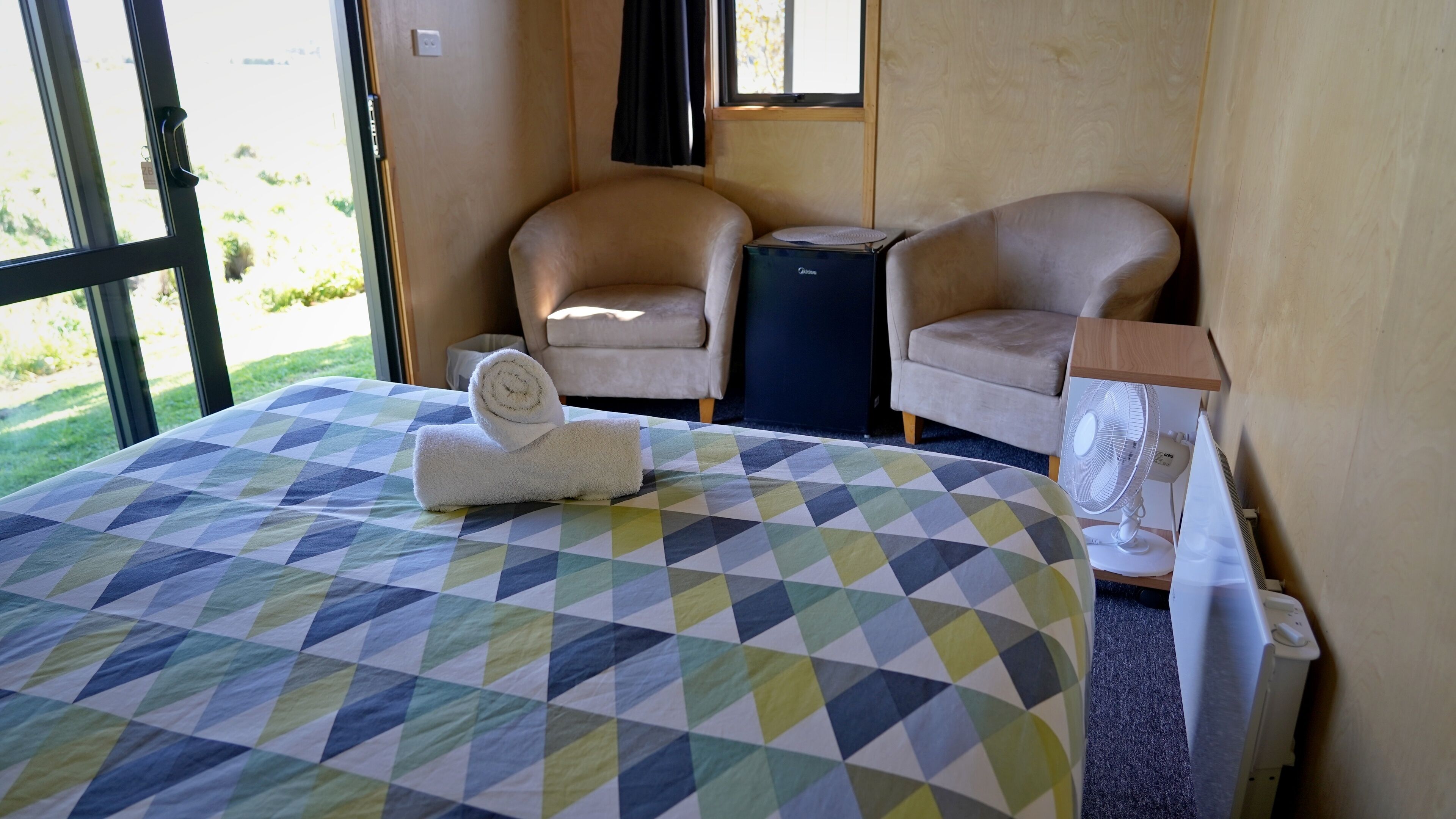 A hotel room with a bed, two chairs, a small table, a fan, and a window with a view of greenery.