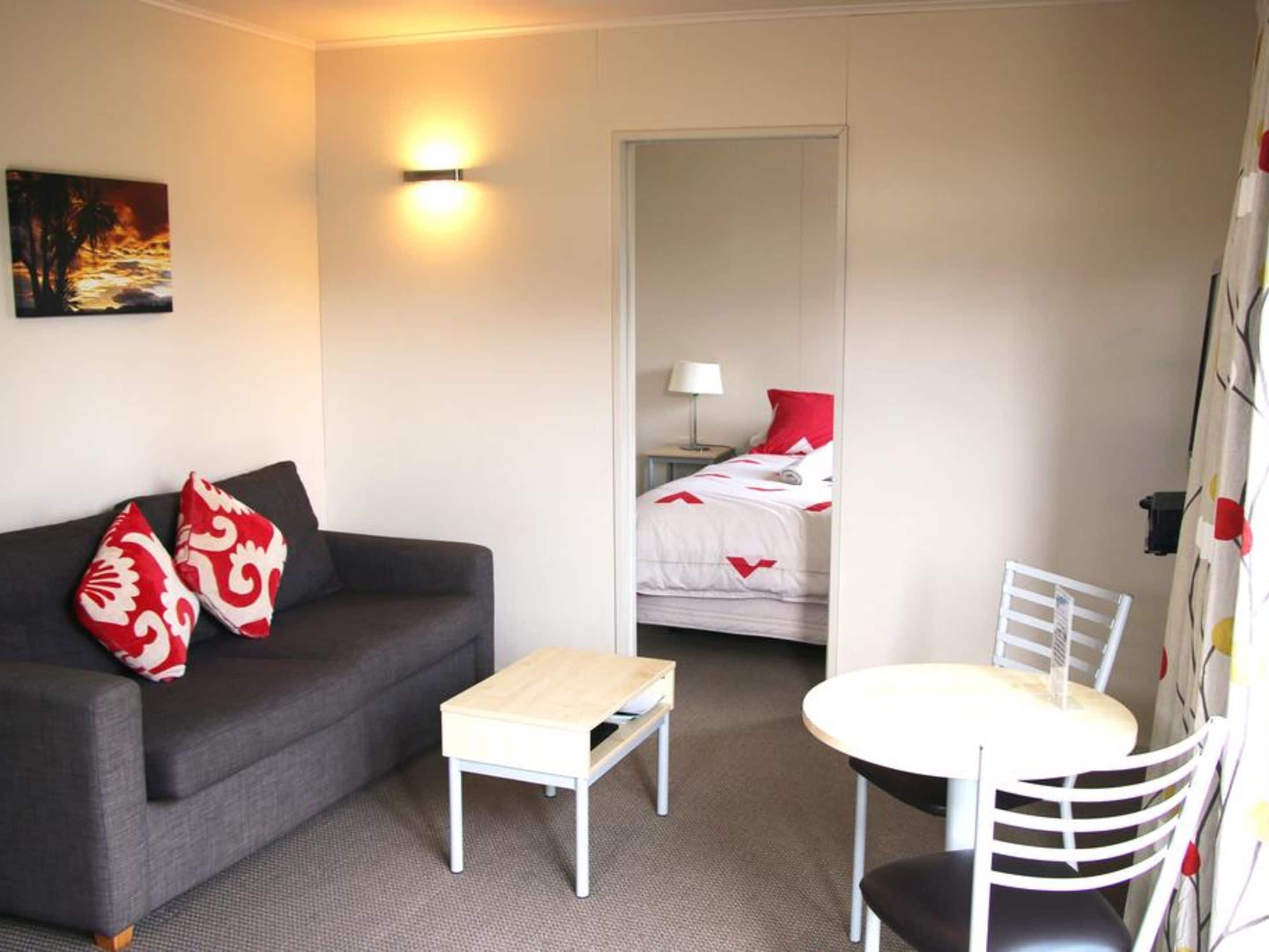 A room with a grey sofa, a small white table, a white chair, a bed with red bedding, and a painting on the wall.