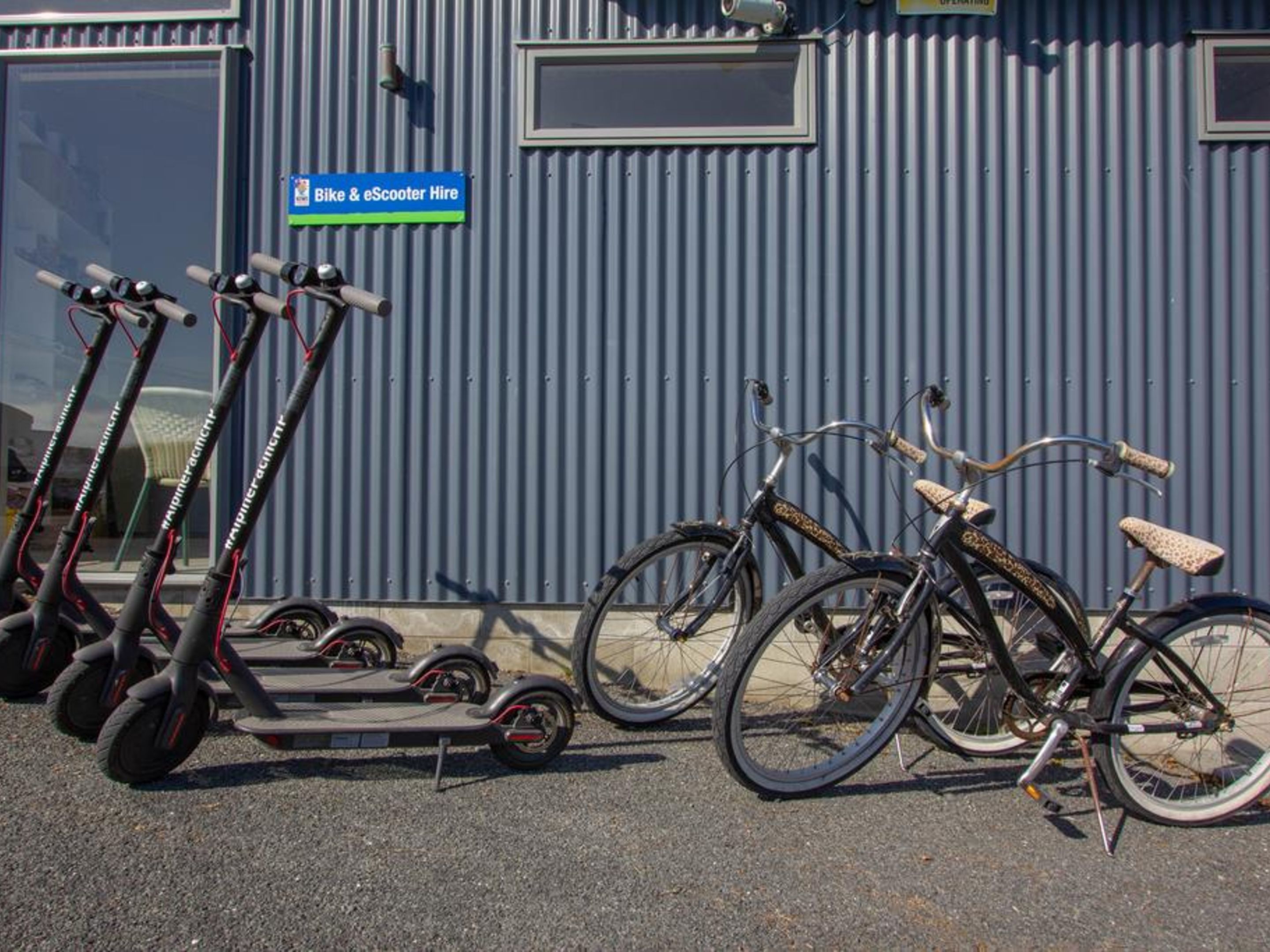 Una fila de scooters eléctricos y bicicletas estacionadas frente a un muro de metal corrugado con un letrero que indica un servicio de alquiler de bicicletas y scooters eléctricos.