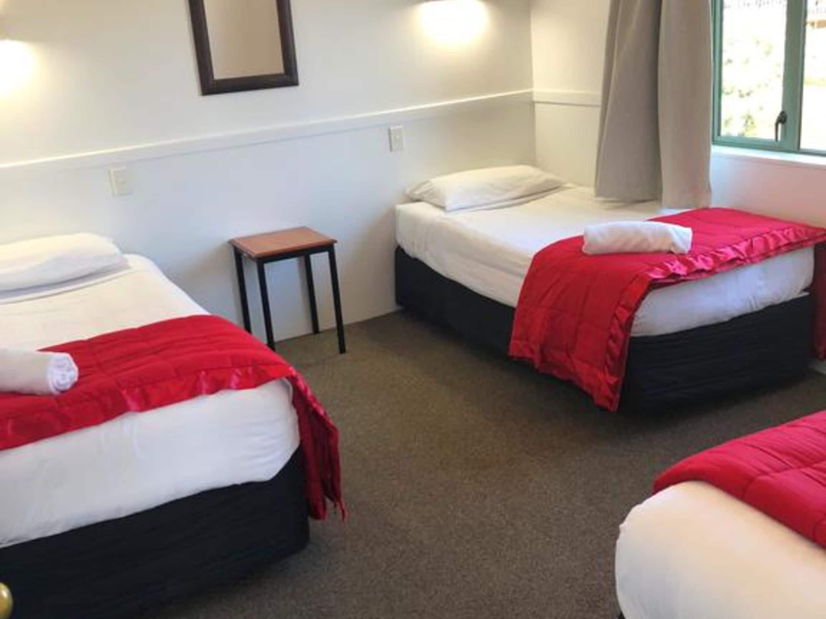 A hotel room with three single beds, each with white bedding and red blankets. There is a small wooden table beside one bed.