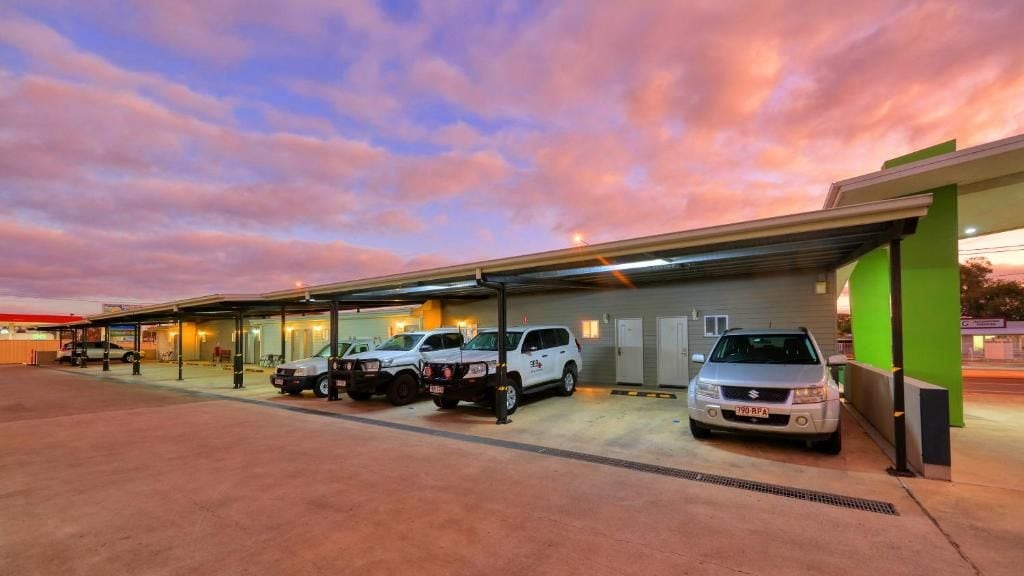 Un estacionamiento con varios vehículos, una estructura cubierta y un edificio con detalles verdes.