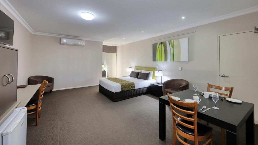 A hotel room with a bed, a dining table with chairs, a refrigerator, and a wall-mounted air conditioner.