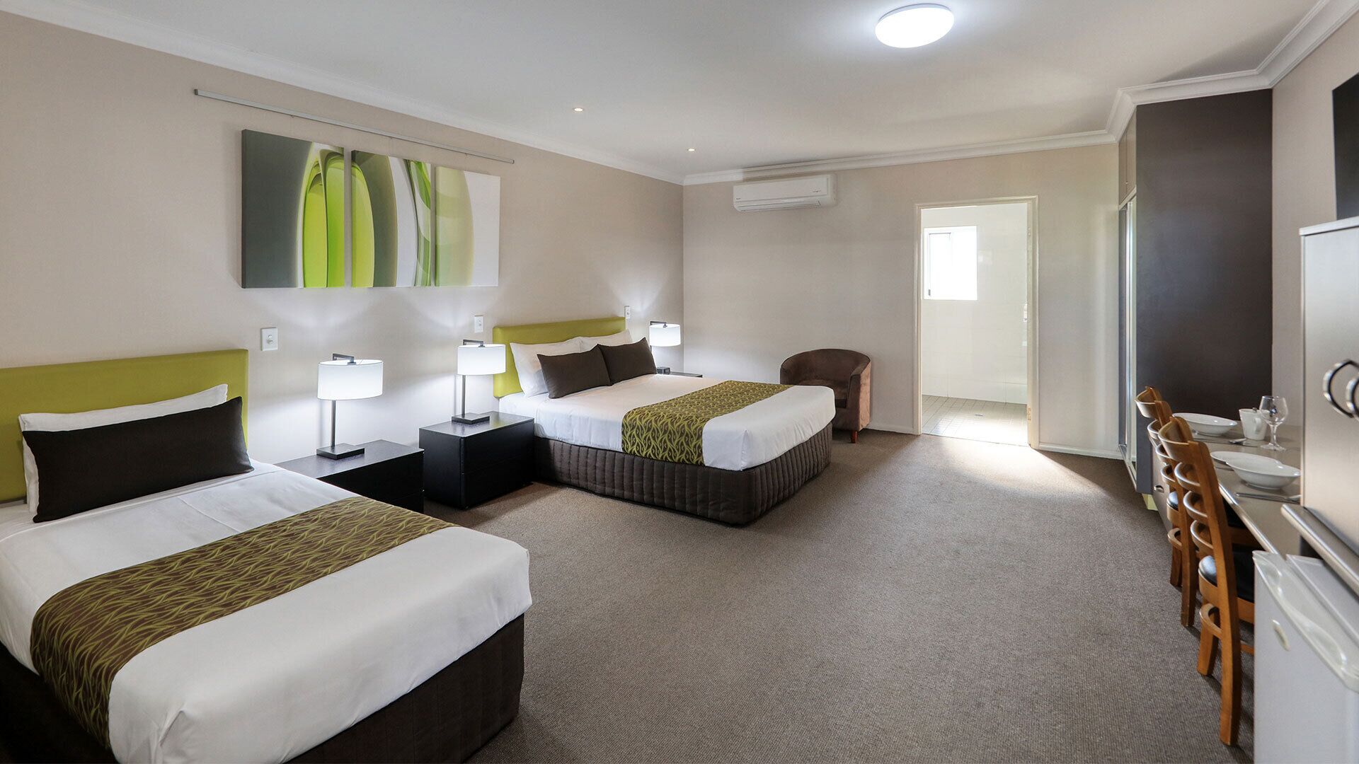 A hotel room with two beds, a chair, a desk with a chair, and a dining area with a refrigerator.