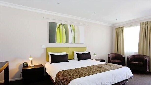 Una habitación de hotel con una cama, una mesita de noche con lámpara, una silla y una ventana con cortinas.