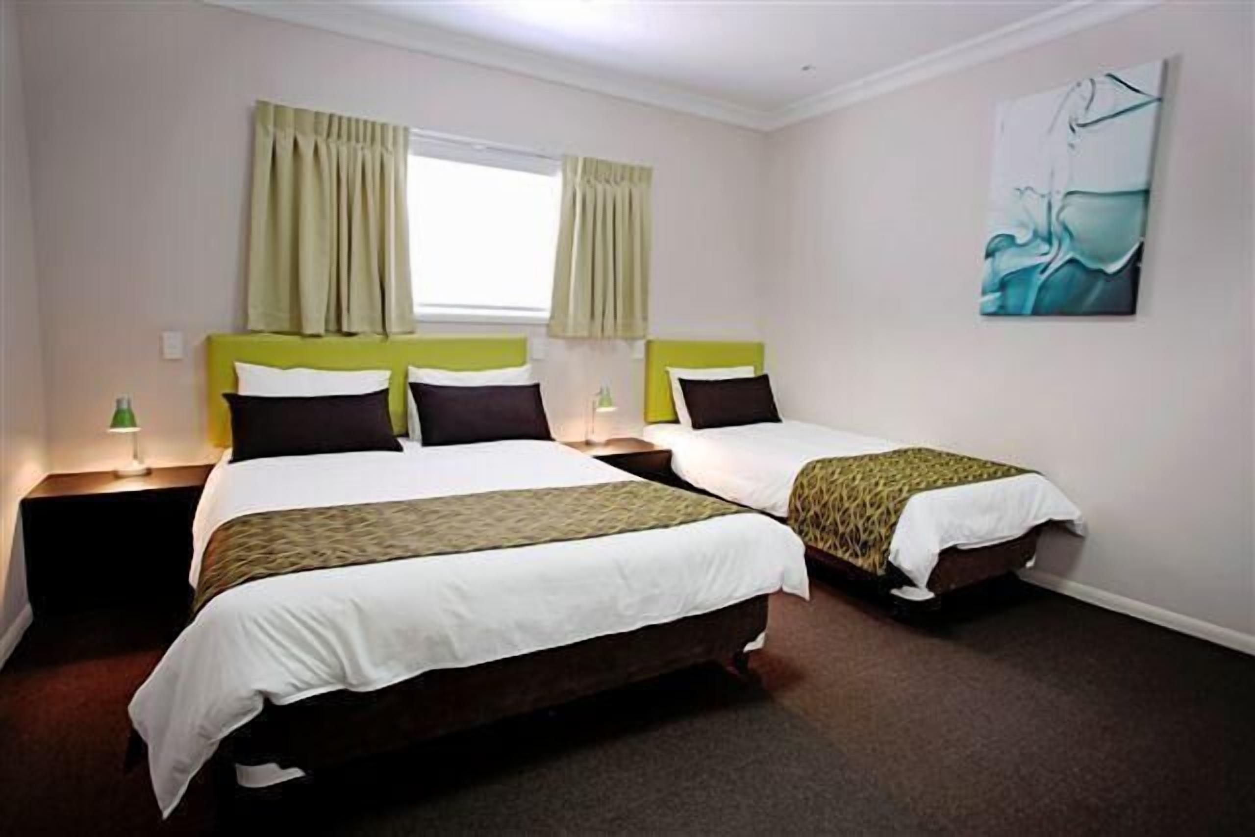 A hotel room with two beds, a window with curtains, a painting on the wall, and a lamp on the nightstand.