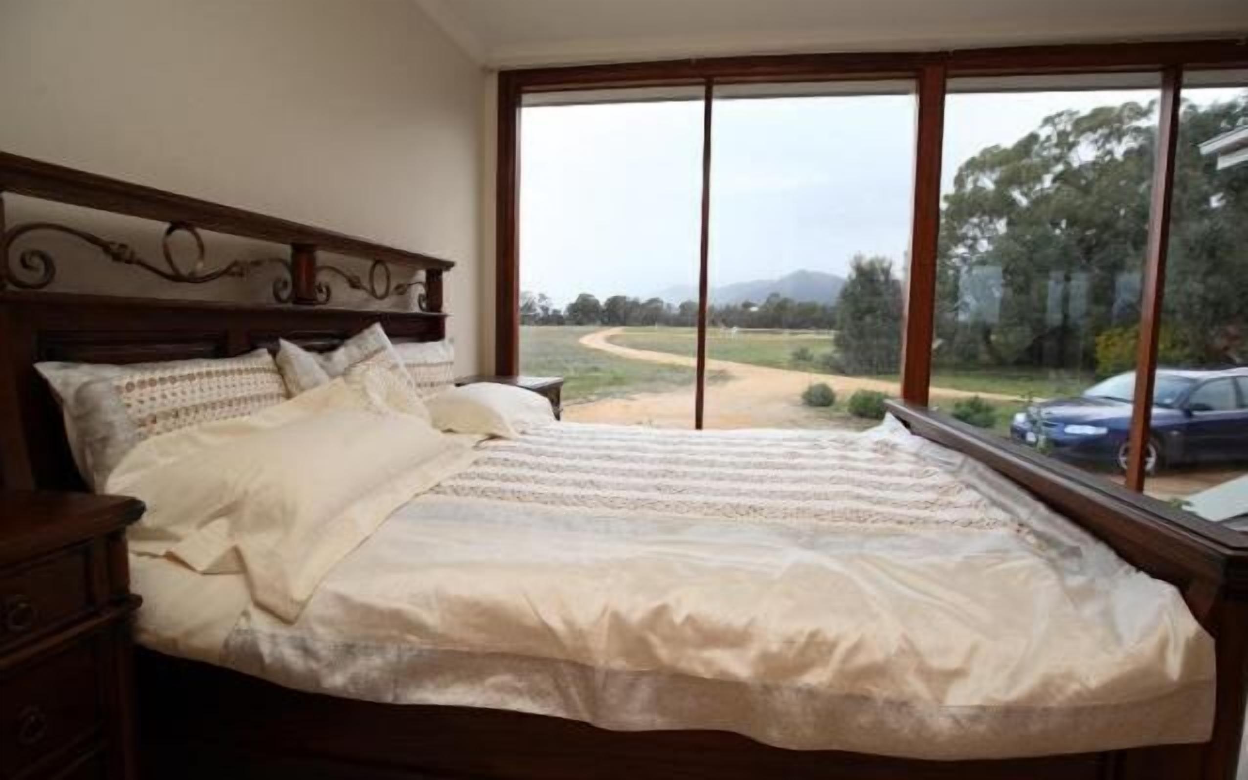 Une chambre à coucher avec un grand lit, une tête de lit en bois et une vue sur un chemin de terre et des arbres, visible par la fenêtre.