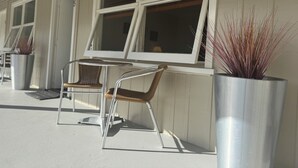 Ein überdachter Außenbereich mit Metallstühlen und einem Tisch, Zimmerpflanzen und großen Fenstern mit Jalousien.