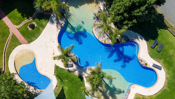Ein Luftbild eines Schwimmbadkomplexes mit mehreren verbundenen Pools, umgeben von gepflegtem Rasen und Palmen.