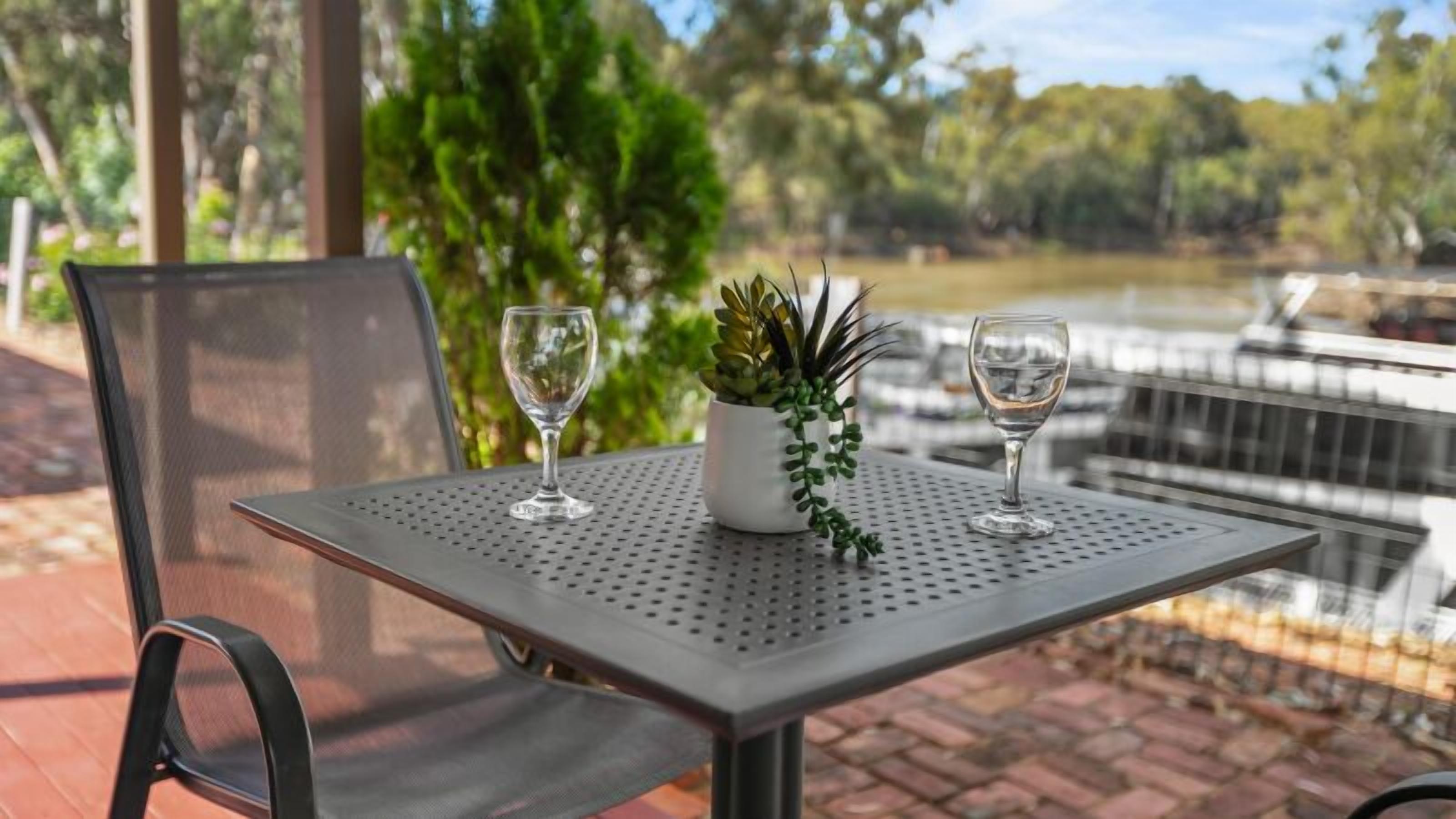 Une table en métal à surface perforée, sur laquelle sont posés deux verres et un pot de fleurs, est installée à l’extérieur. On aperçoit, en arrière-plan, des arbres et une étendue d’eau.