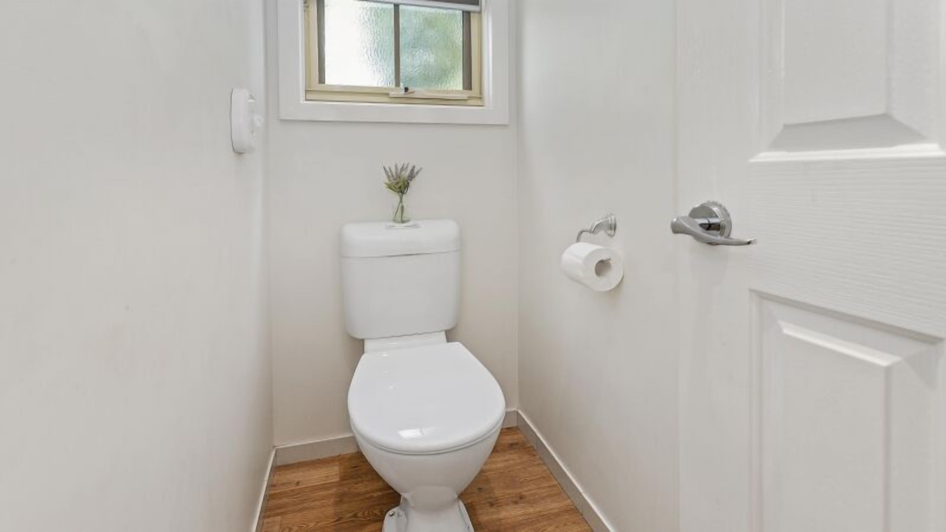 A small, white bathroom with a wooden floor, a toilet, a window, and a door.